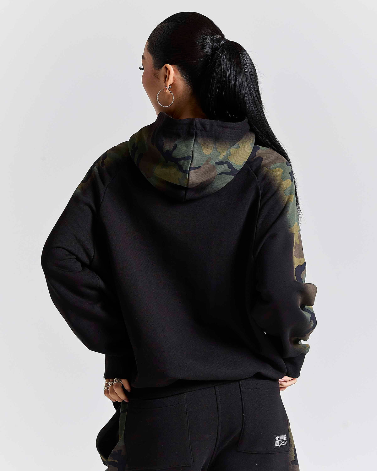A woman shown from the back wearing a black hoodie with a green and brown camouflage pattern on the hood and upper sleeves, paired with black sweatpants against a light gray studio background.