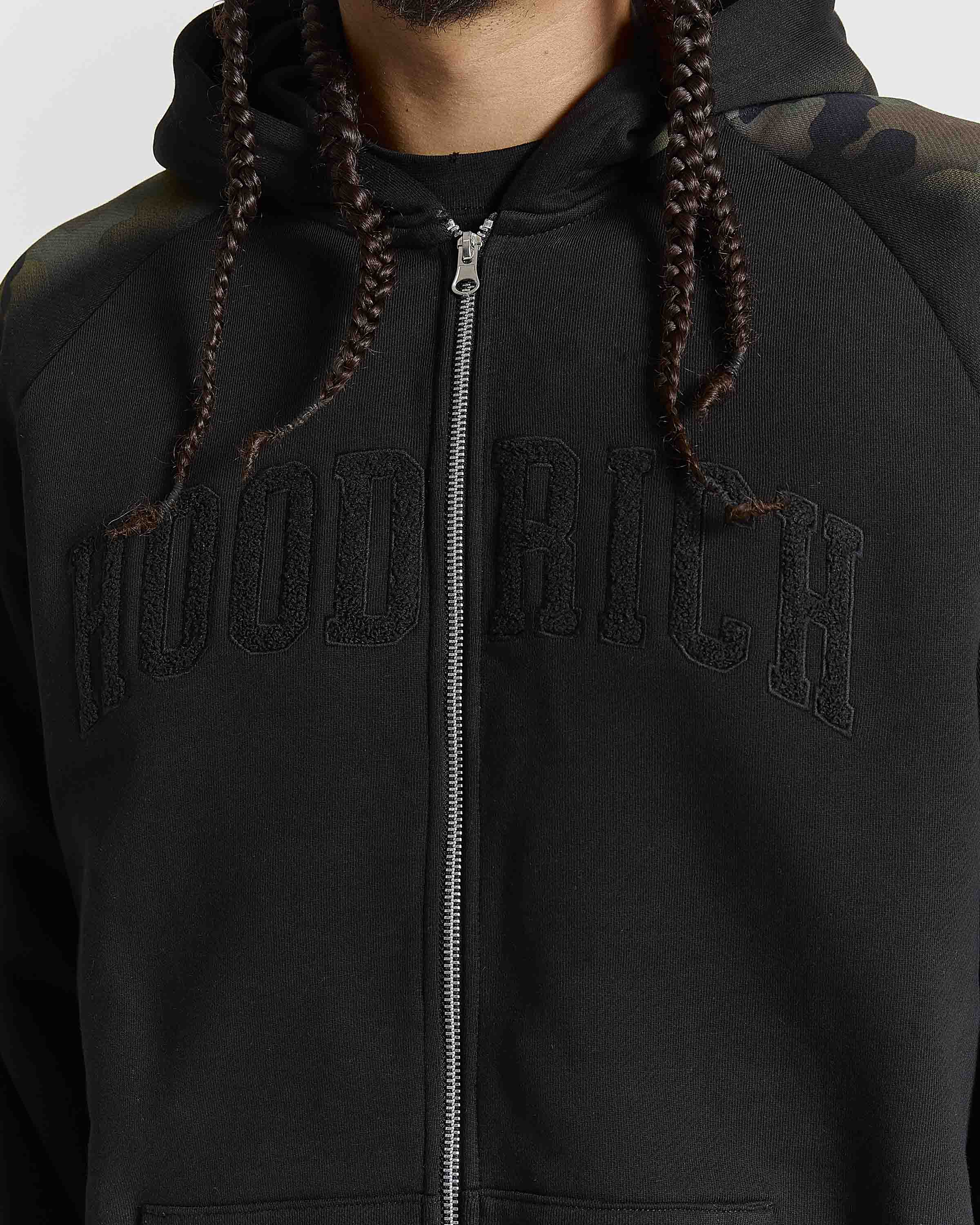 A close-up of a person wearing a black zip-up hoodie with 'HOODRICH' embroidered in tonal black across the chest. The hoodie has a silver zipper, black drawstrings, and a subtle camouflage pattern on the right shoulder.