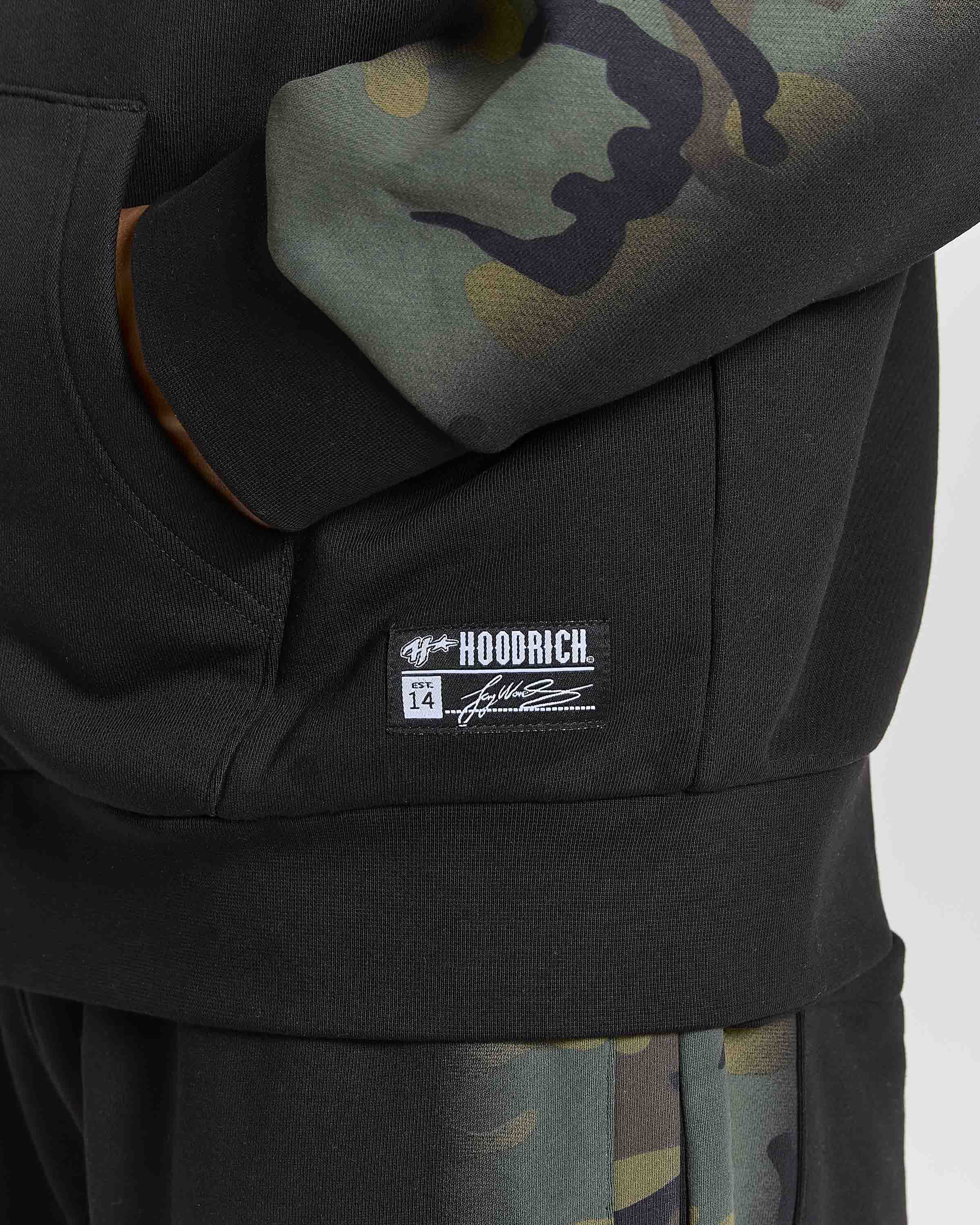 Close-up of a black hoodie with a kangaroo pocket, featuring a white HOODRICH brand patch on the pocket and a green camouflage pattern on the sleeve.