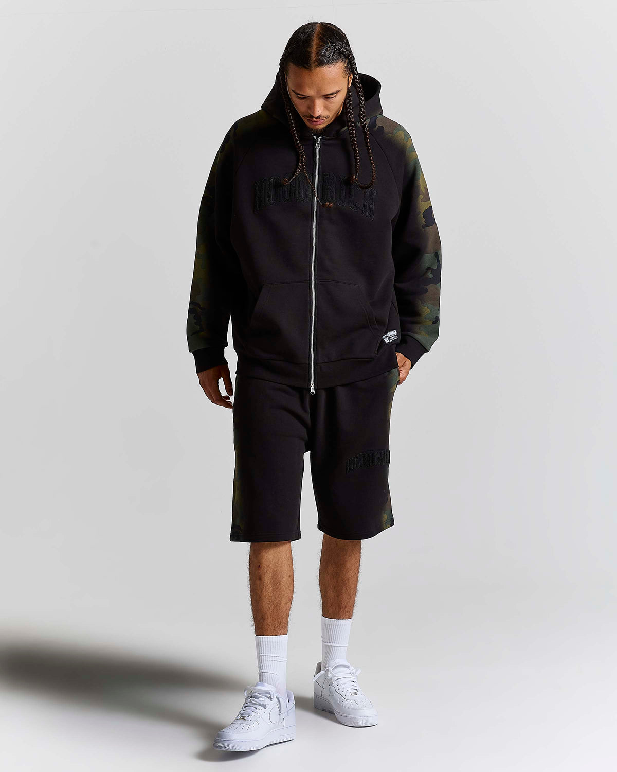 A man modeling a black zip-up hoodie with dark green camouflage sleeves and hood, matching black shorts, white socks, and white sneakers, shot against a light gray studio background.