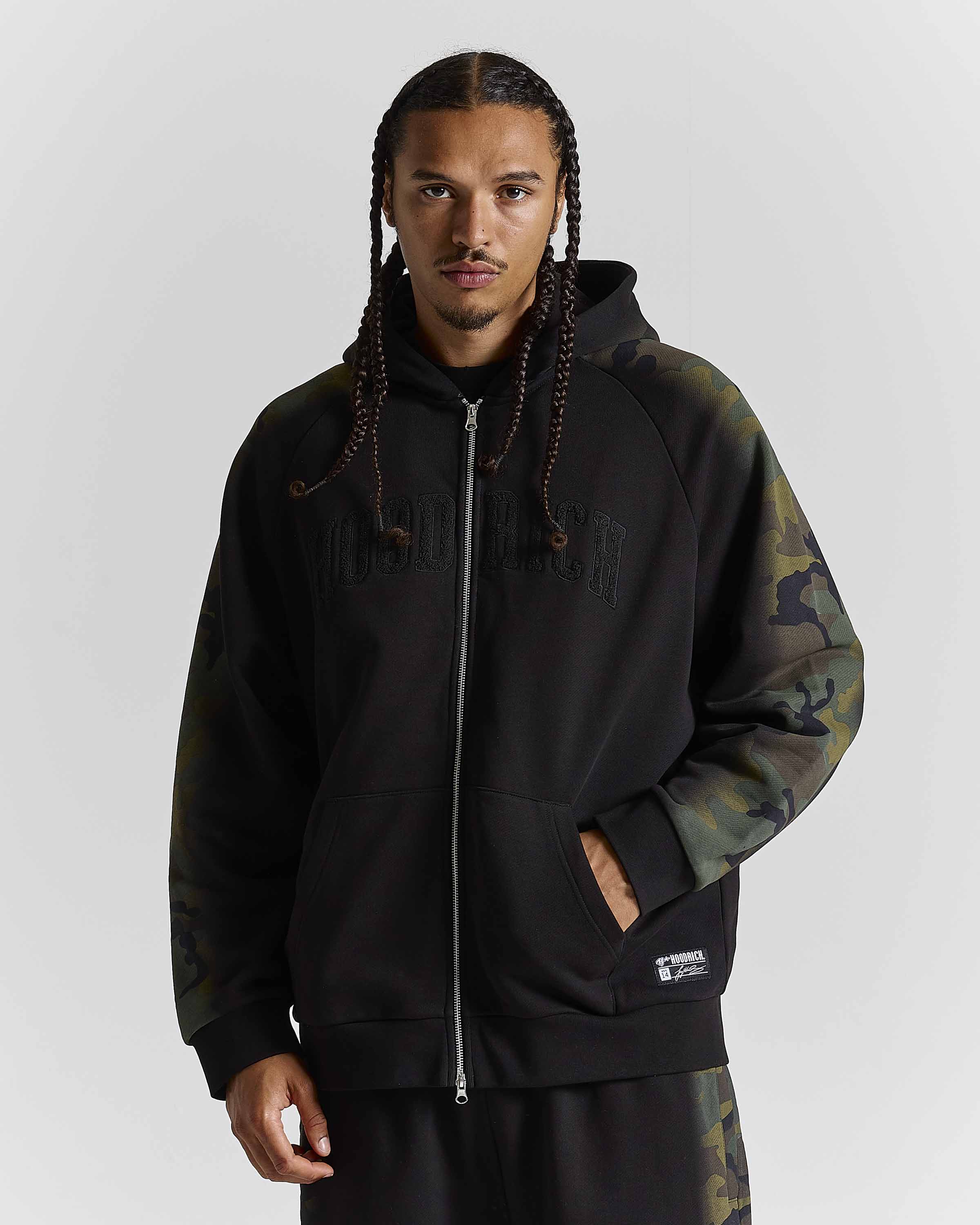 A man wearing a black zip-up hoodie with dark camouflage sleeves and a matching camouflage sweatpants, photographed against a neutral gray studio background.