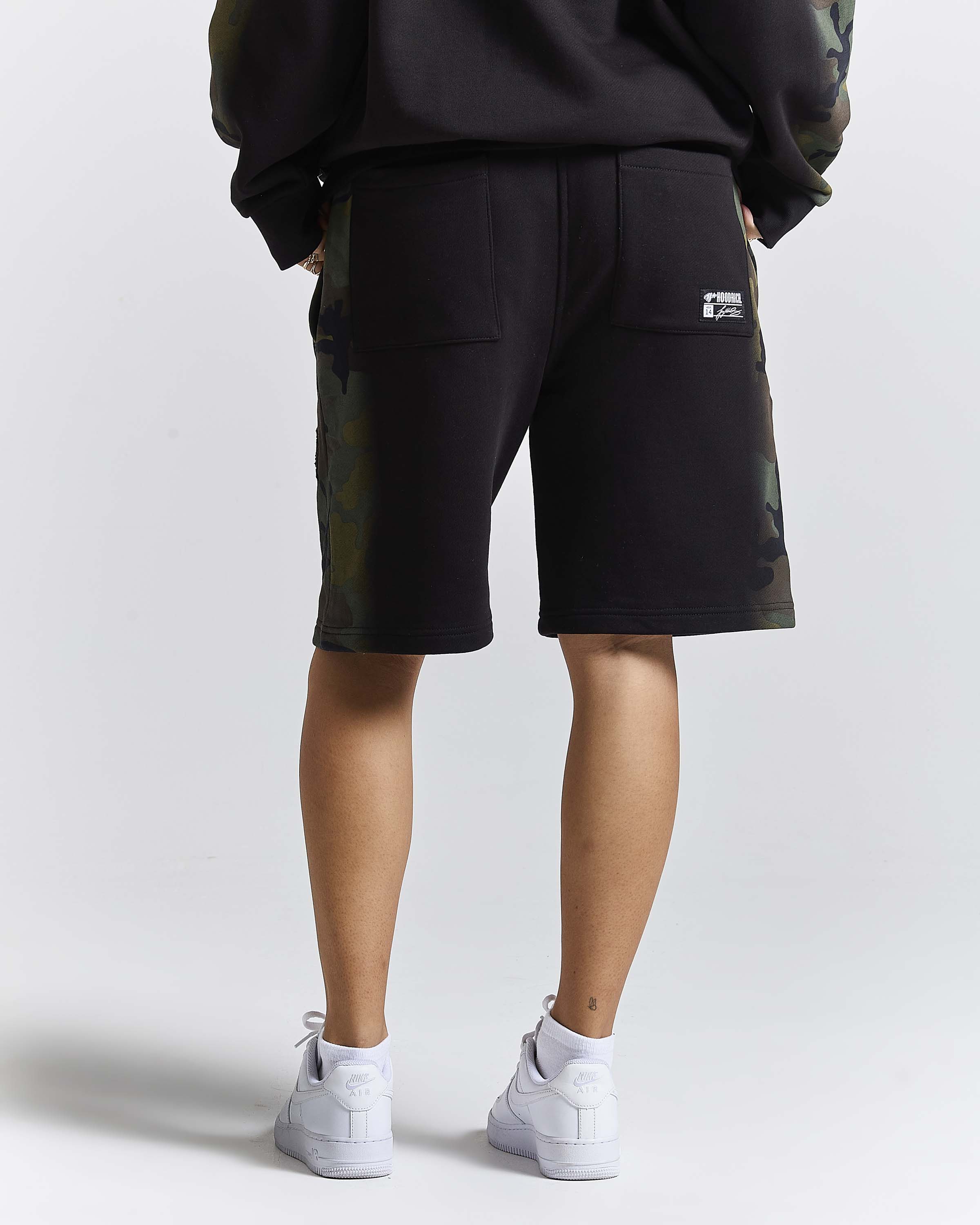 A person shown from the back wearing black shorts with camouflage side panels, a black sweatshirt, and white sneakers against a light gray studio background.