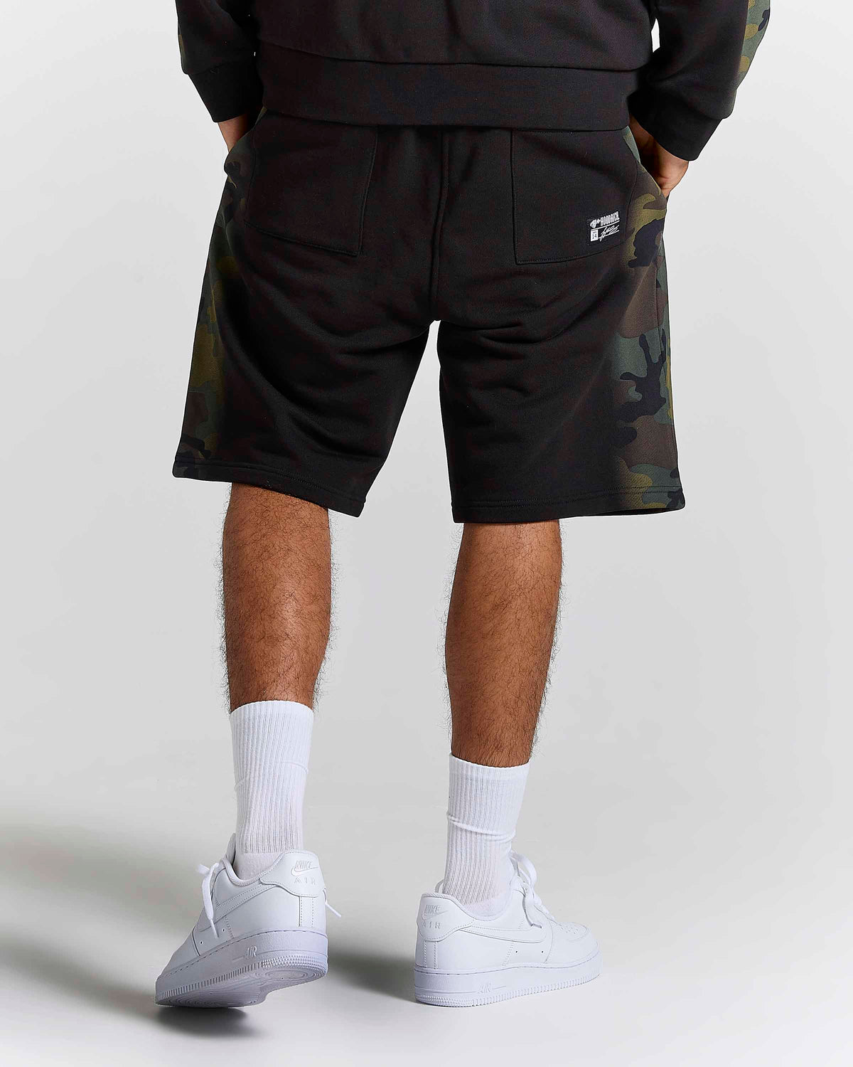 Rear view of a person wearing black athletic shorts with green and brown camouflage side panels, white crew socks, and white low-top sneakers against a light gray studio background.