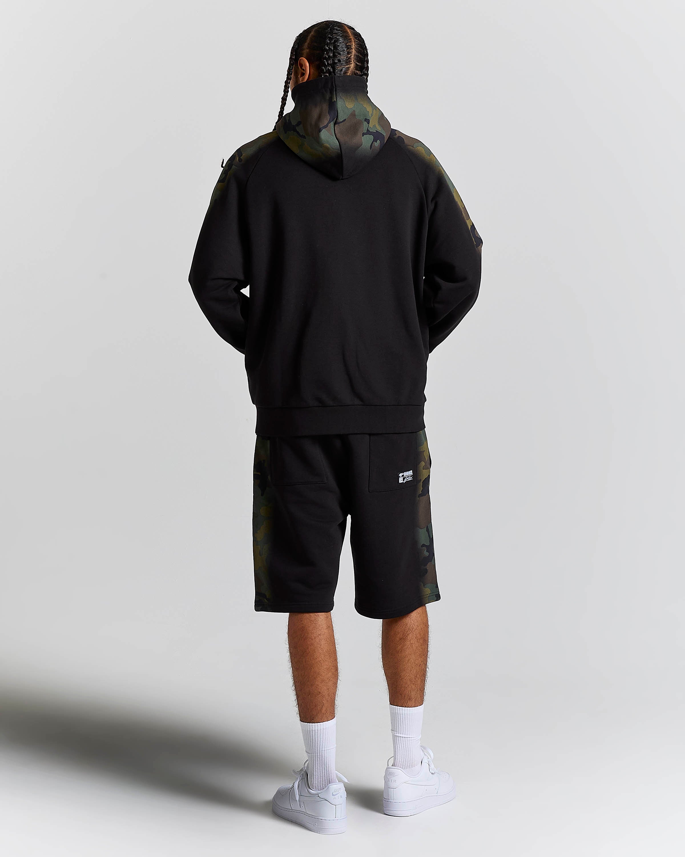 A model shown from the back wearing a black hoodie with camouflage pattern details on the hood and sleeves, matching camouflage shorts, white socks, and white sneakers, set against a plain light gray studio background.