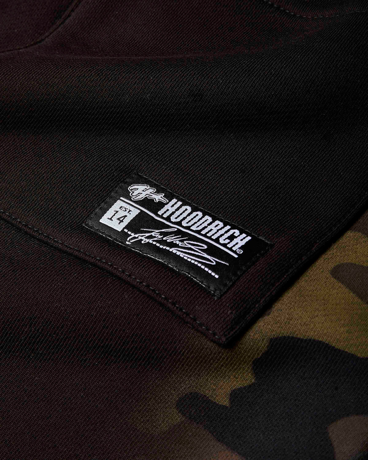 Close-up of a black HOODRICH brand patch sewn onto a dark brown and black camouflage hoodie.