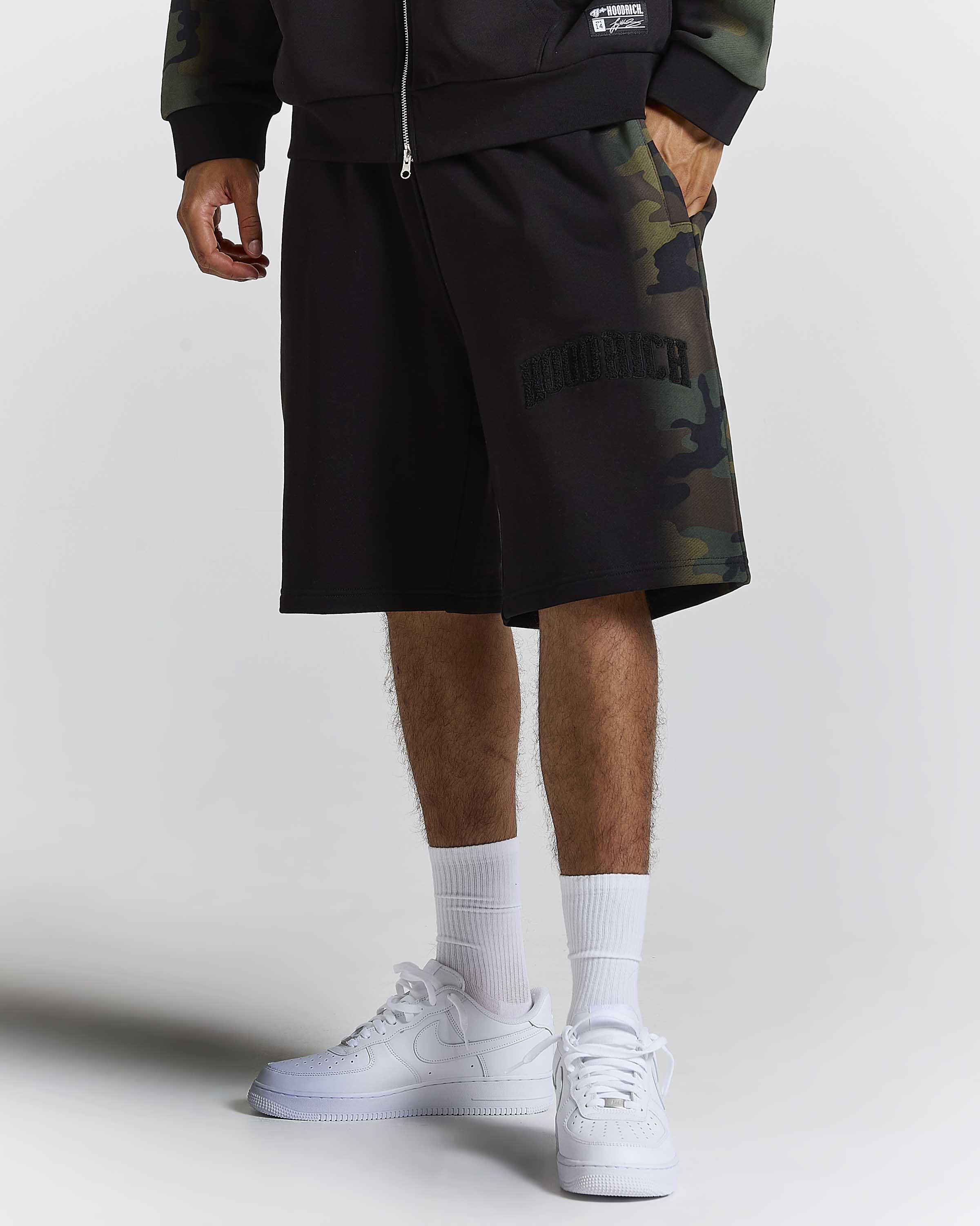 A person wearing black camouflage athletic shorts with a tonal 'HOODRICH' logo, a matching zip-up sweatshirt, white socks, and white sneakers, photographed against a light gray studio background.