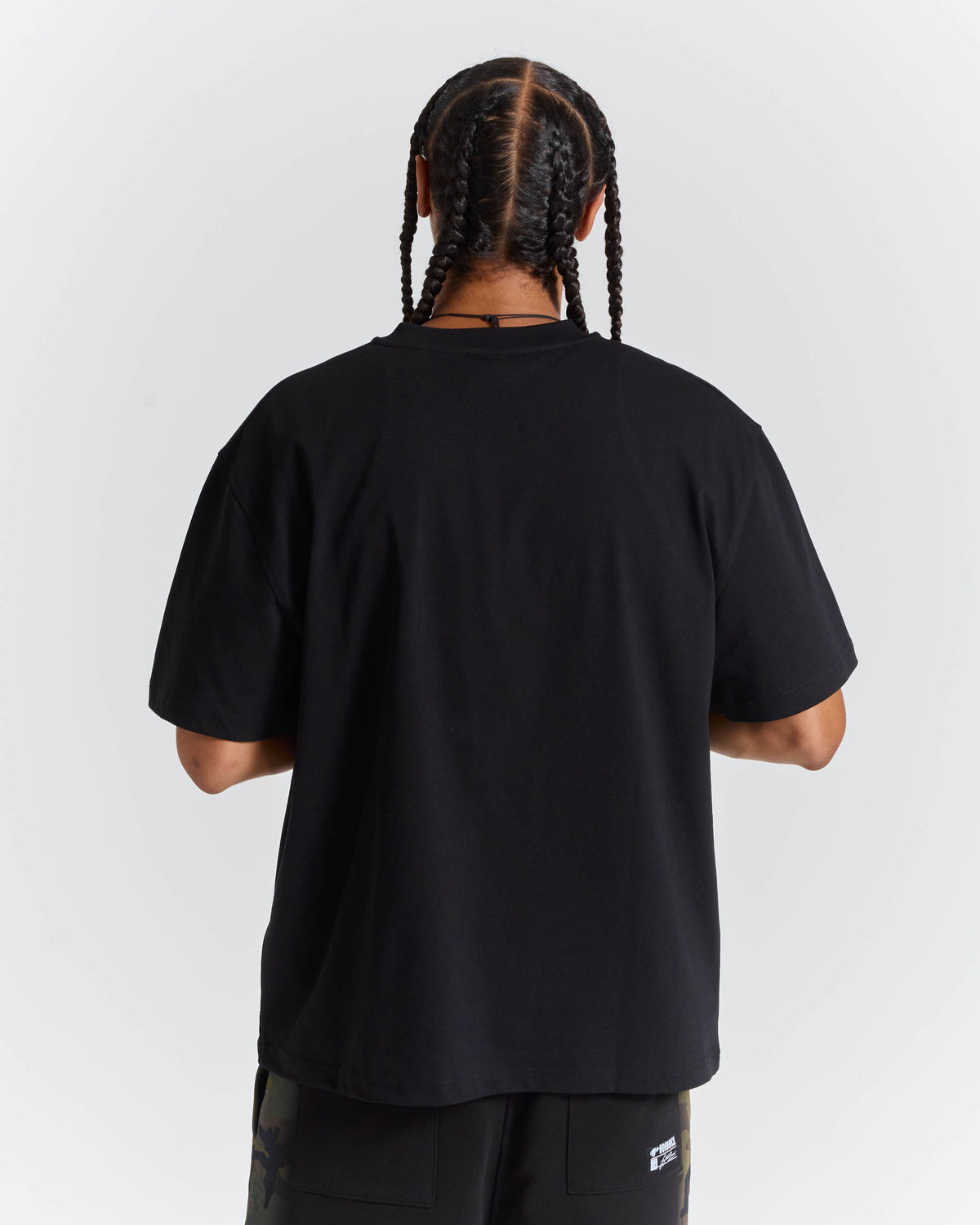 A person shown from the back wearing an oversized black short-sleeve T-shirt and dark pants against a light gray studio background.