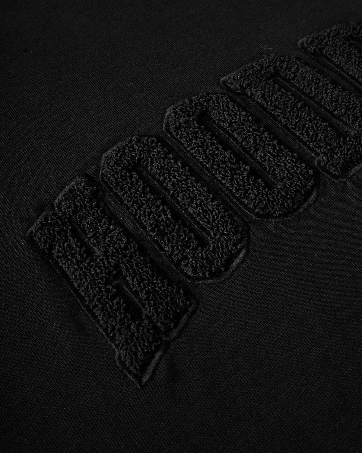 Close-up of a black textured fabric with large raised embroidered letters reading 'HOODRICH' in black-on-black thread.