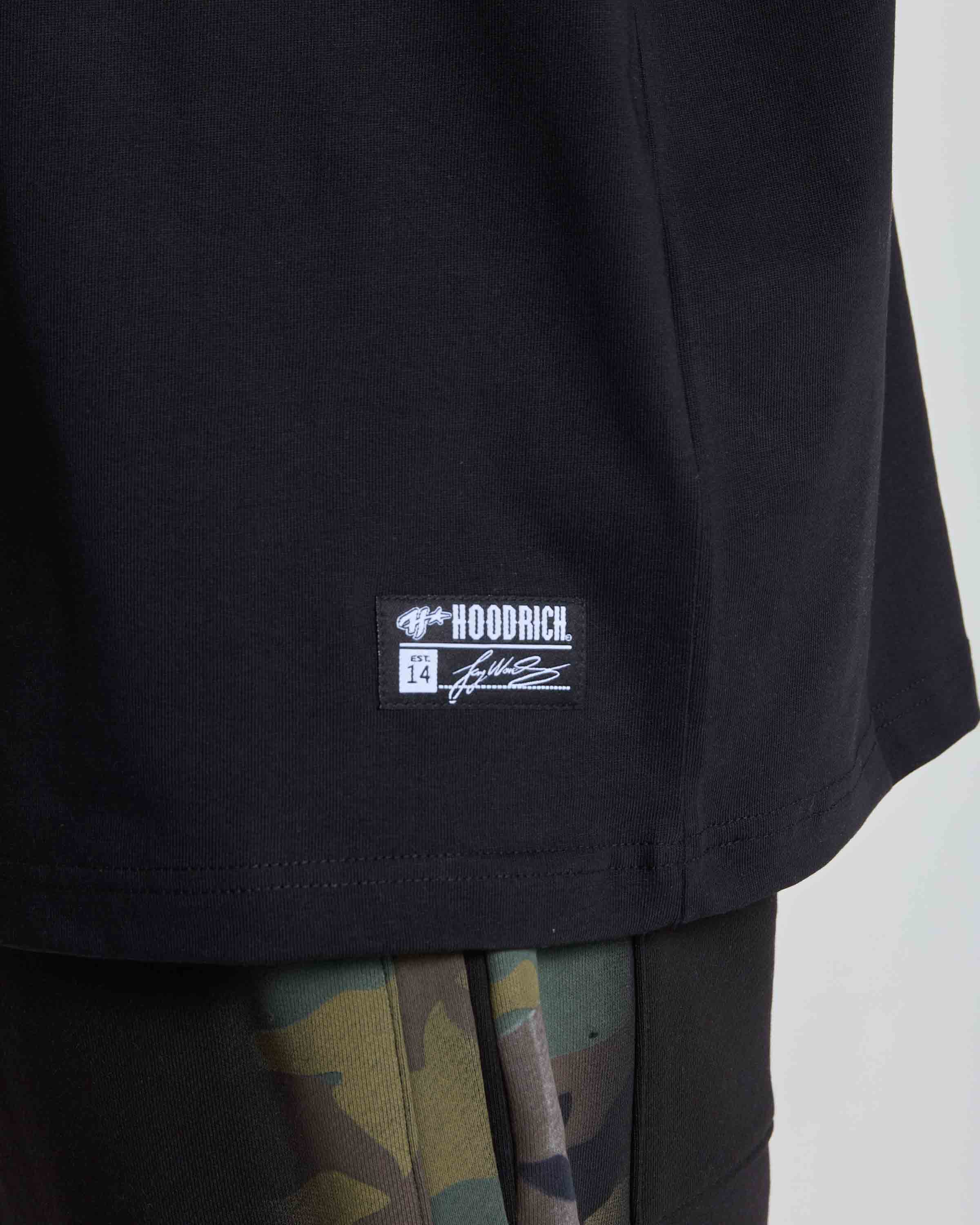Close-up of a black HOODRICH t-shirt showing the brand patch on the lower front, worn over camouflage pants.
