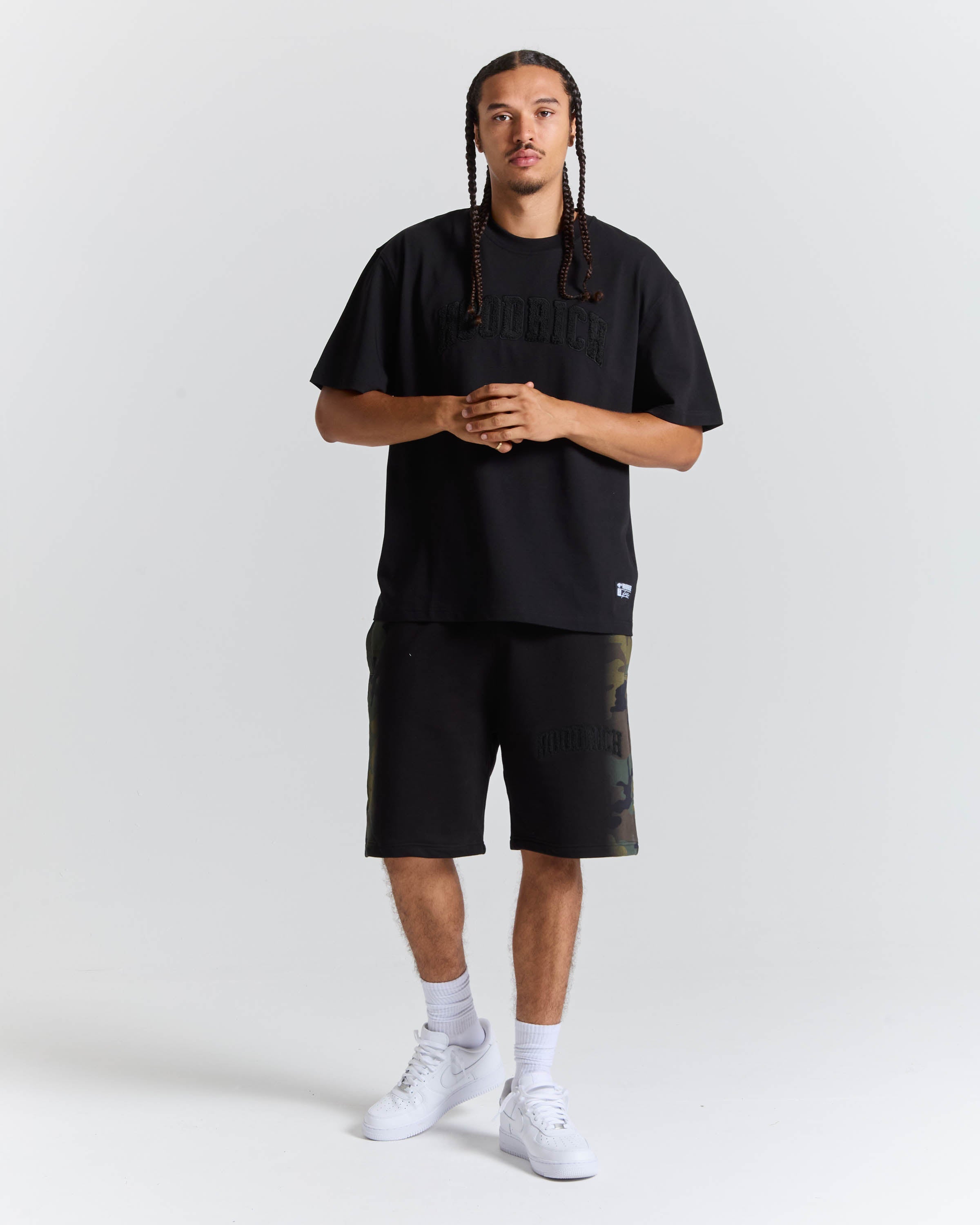A man modeling a black short-sleeve t-shirt and matching black shorts with subtle camouflage details on the side, paired with white socks and white sneakers, against a light gray studio background.