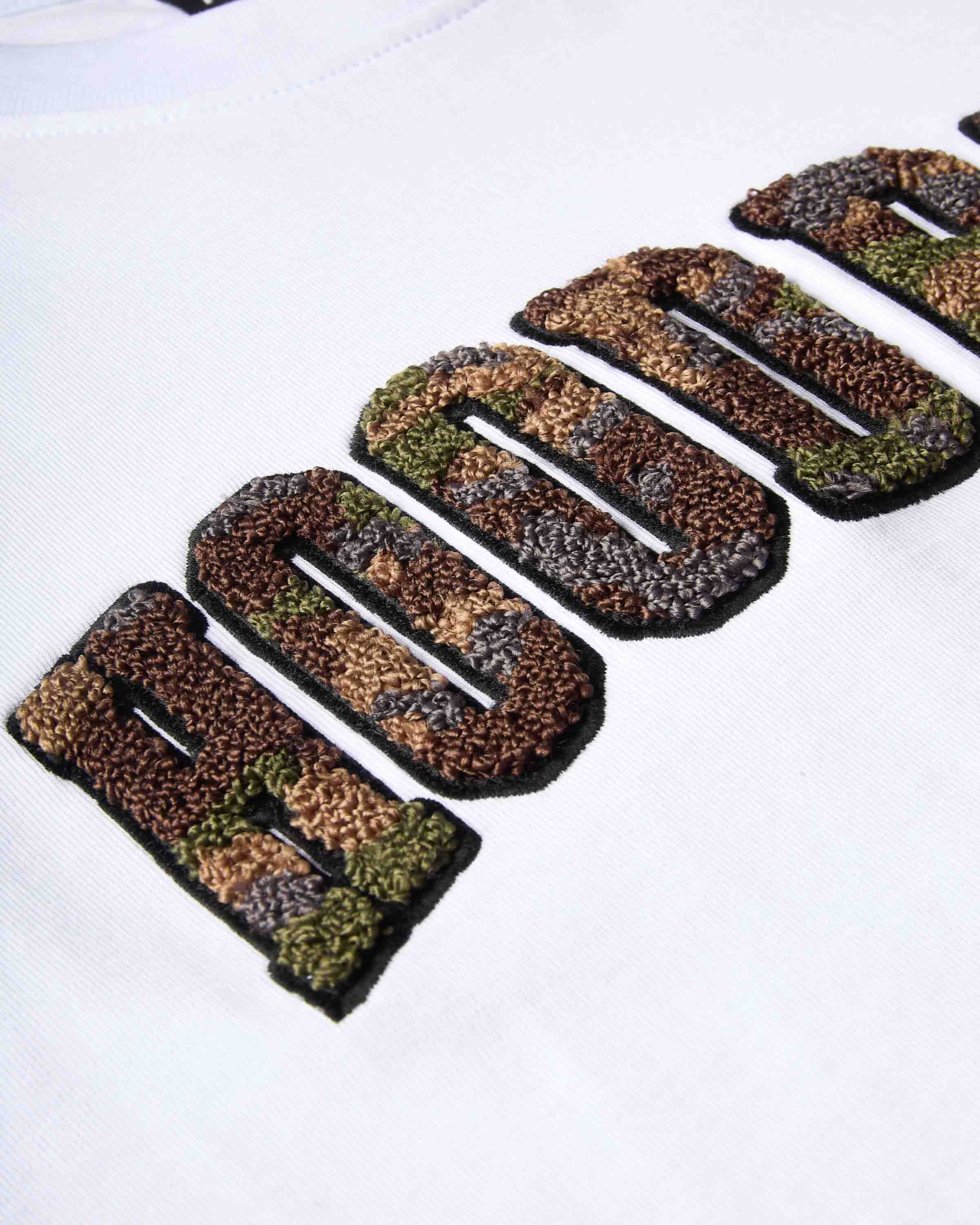 Close-up of a white t-shirt featuring a large, textured embroidered graphic of 'HOODRICH' in a camouflage pattern with black outlines.