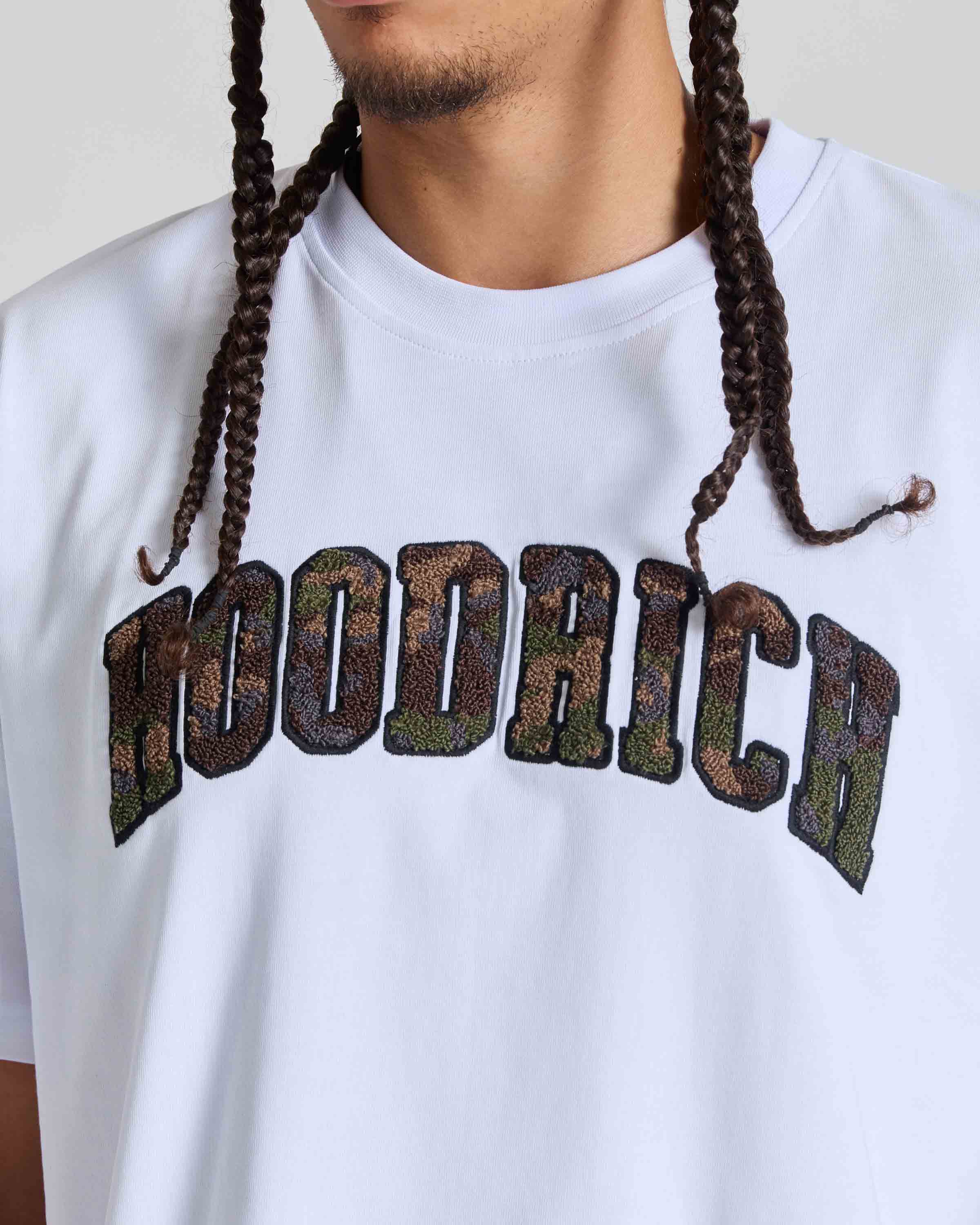 A close-up of a person wearing a white crewneck t-shirt with the brand name 'HOODRICH' printed across the chest in large, arched letters filled with a camouflage chenille pattern.