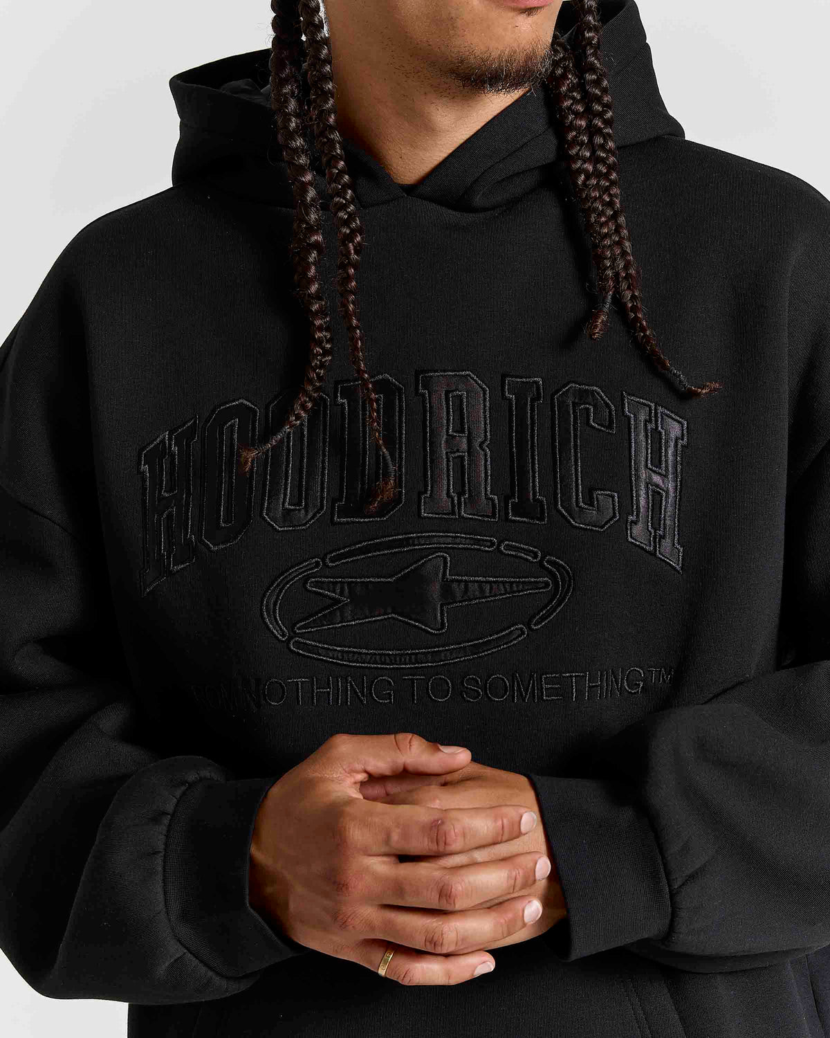 A close-up of a person wearing a black pullover hoodie with tonal black graphic text reading 'HOODRICH' and a star logo on the chest, against a light gray background.
