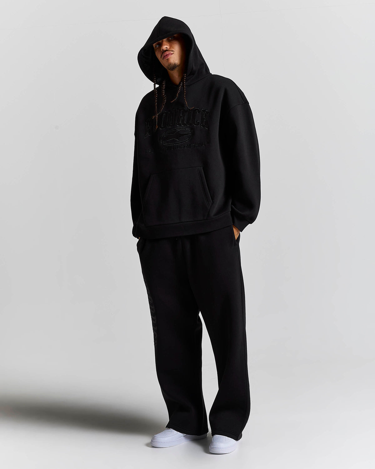 A model wearing a black oversized hoodie with a tonal black graphic on the chest, matching black sweatpants, and white sneakers, standing against a light gray studio background.