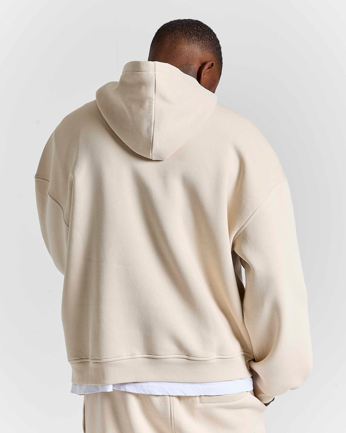A man viewed from behind wearing a light beige oversized hoodie and matching sweatpants against a plain light gray studio background.