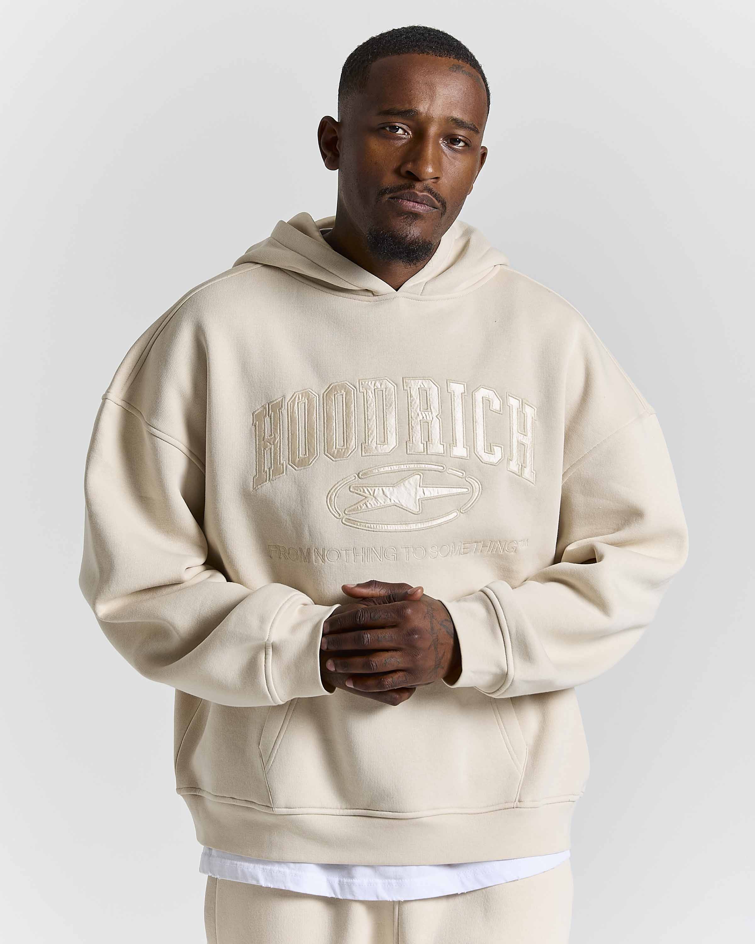 A man wearing an oversized beige hoodie with a tonal 'HOODRICH' logo and fish graphic on the chest, paired with matching beige pants, against a light gray studio background.