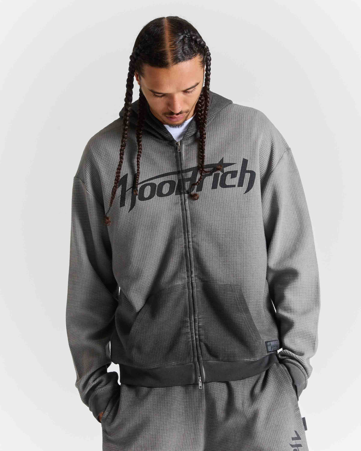 A man wearing a gray zip-up hoodie with a black 'hoodrich' logo across the chest, paired with matching gray sweatpants, photographed against a light gray studio background.