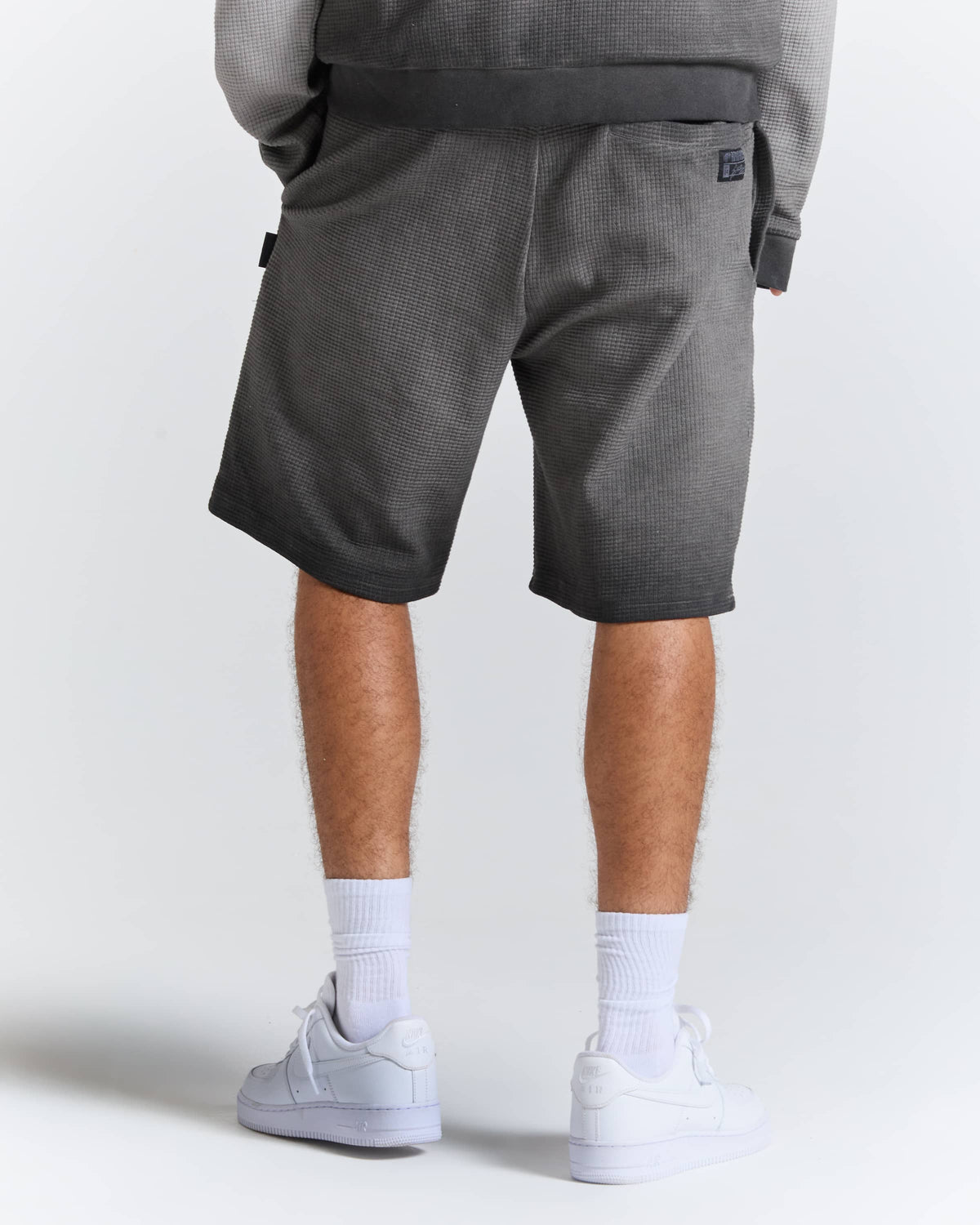 A person shown from the back wearing dark gray textured shorts, a matching gray sweatshirt, white socks, and white sneakers against a light gray studio background.