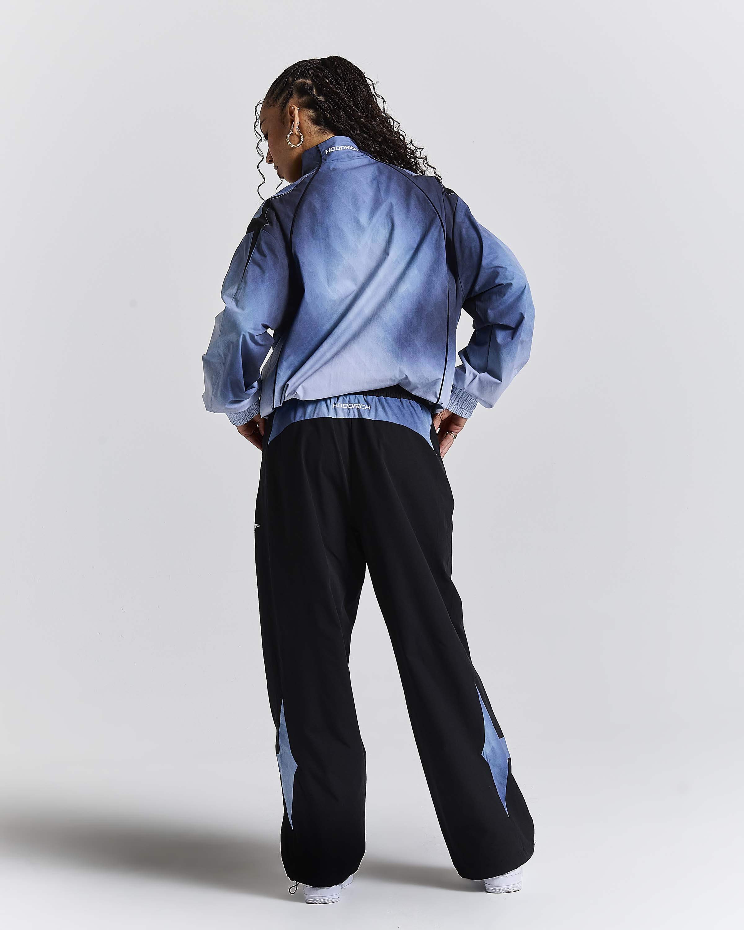 A model is shown from behind wearing a blue tie-dye bomber jacket and black wide-leg sweatpants with blue star accents on the lower legs, set against a plain light gray studio background.