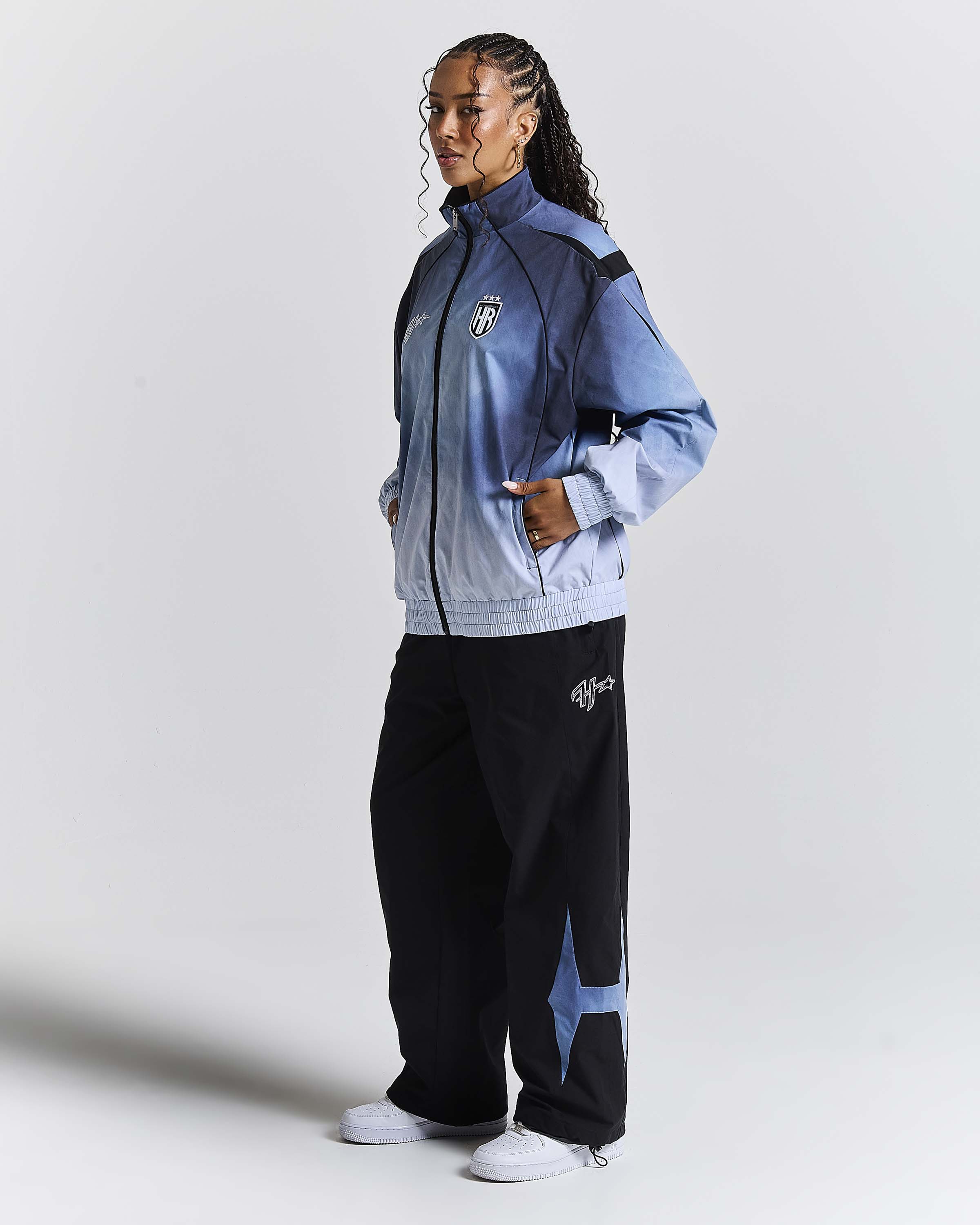 A model wearing a light blue and navy gradient zip-up jacket with black and navy wide-leg sweatpants, standing against a light gray studio background.