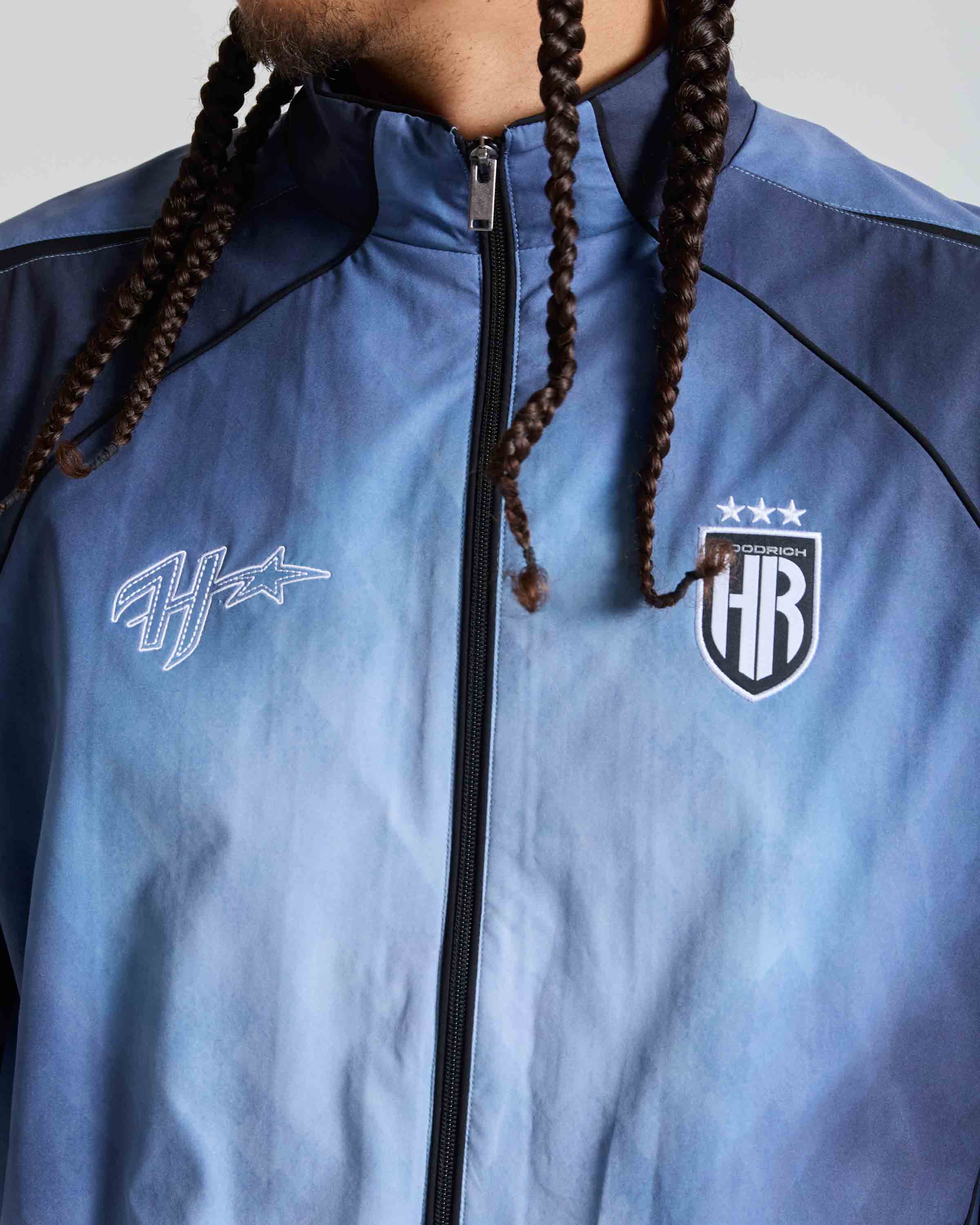 A close-up of a person wearing a light blue zip-up jacket with black piping, featuring the white 'Haze' script logo on the left chest and the 'HRA' shield crest with three stars on the right chest. The person has dark brown braided hair visible at the collar.