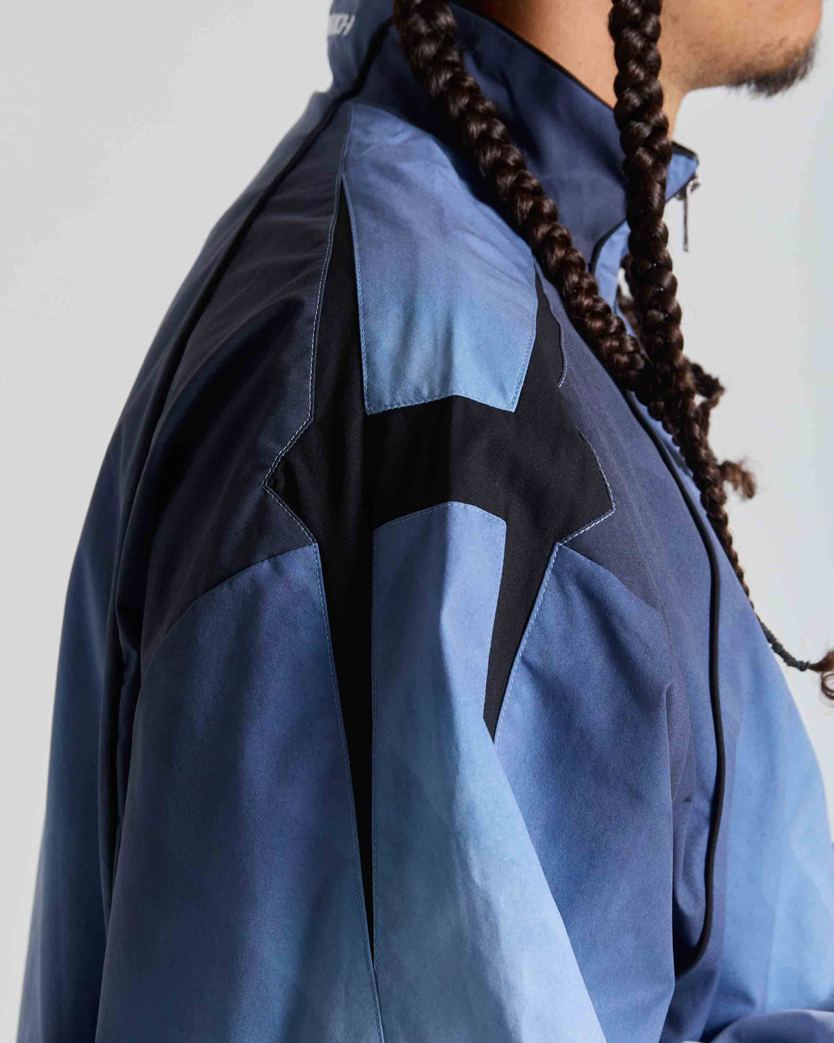 Close-up of a person wearing a blue and black windbreaker jacket, showing the side profile and shoulder details against a light gray background.
