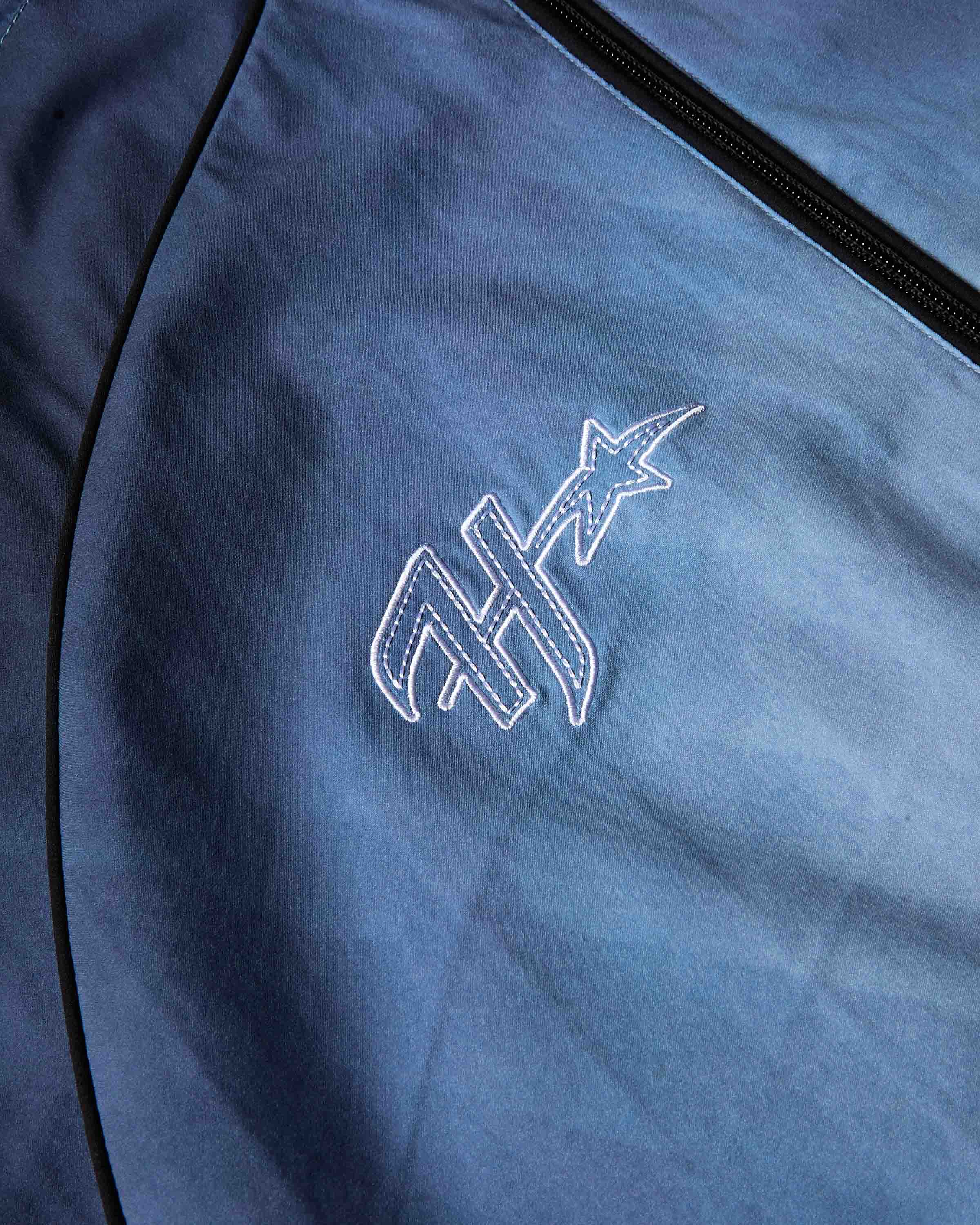 Close-up of a blue zippered garment or bag with black piping and a white embroidered stylized logo featuring a star.