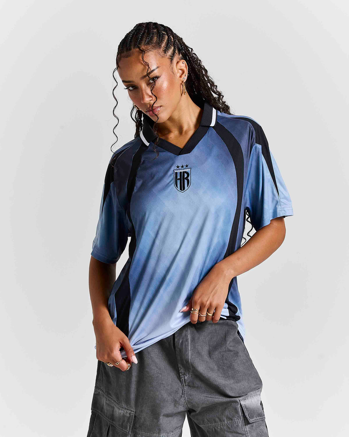 A woman modeling a short-sleeve athletic jersey with a blue-to-light-blue gradient, black side panels, and a black crest on the chest, paired with gray cargo pants against a light gray studio background.