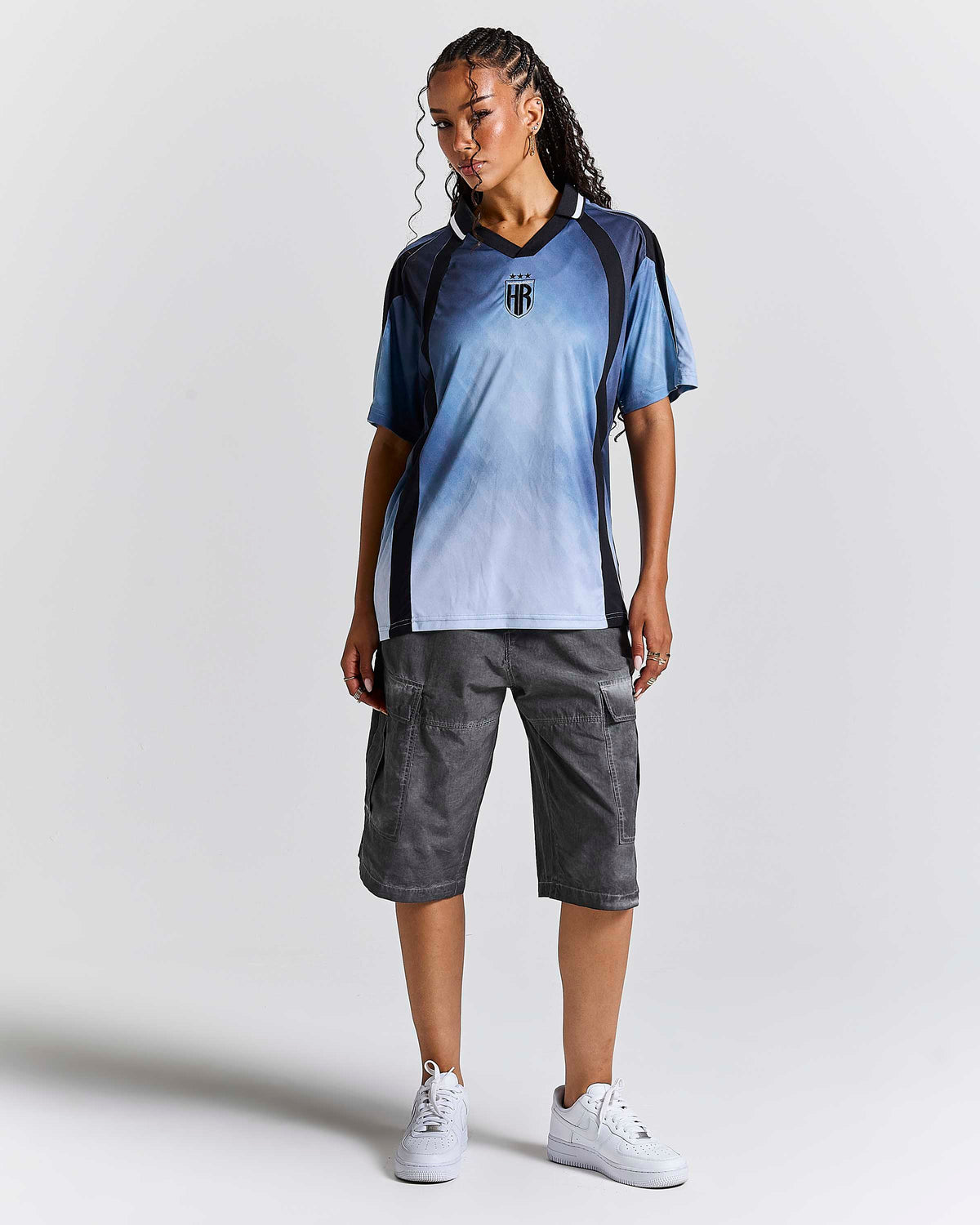 A woman modeling a blue and white gradient soccer jersey with black shoulder panels, paired with dark gray cargo shorts and white sneakers, against a light gray studio background.