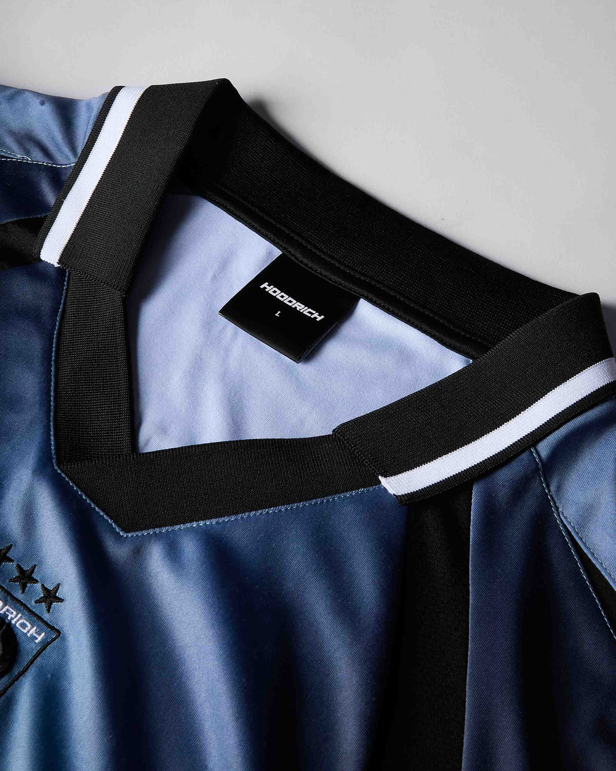 Close-up of a blue V-neck soccer jersey with a black collar trim and white stripes, featuring a black HOODRICH neck label.