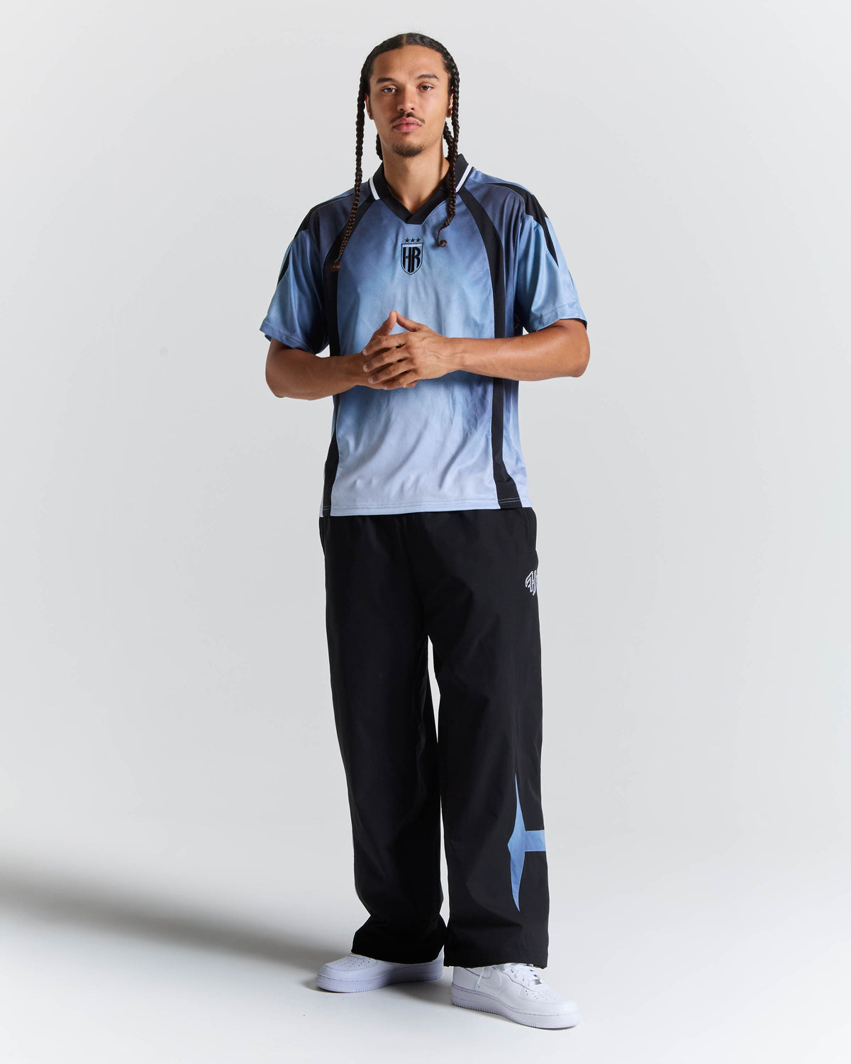 A man modeling a light blue and navy athletic polo shirt with black and navy wide-leg sweatpants, standing against a light gray studio background.