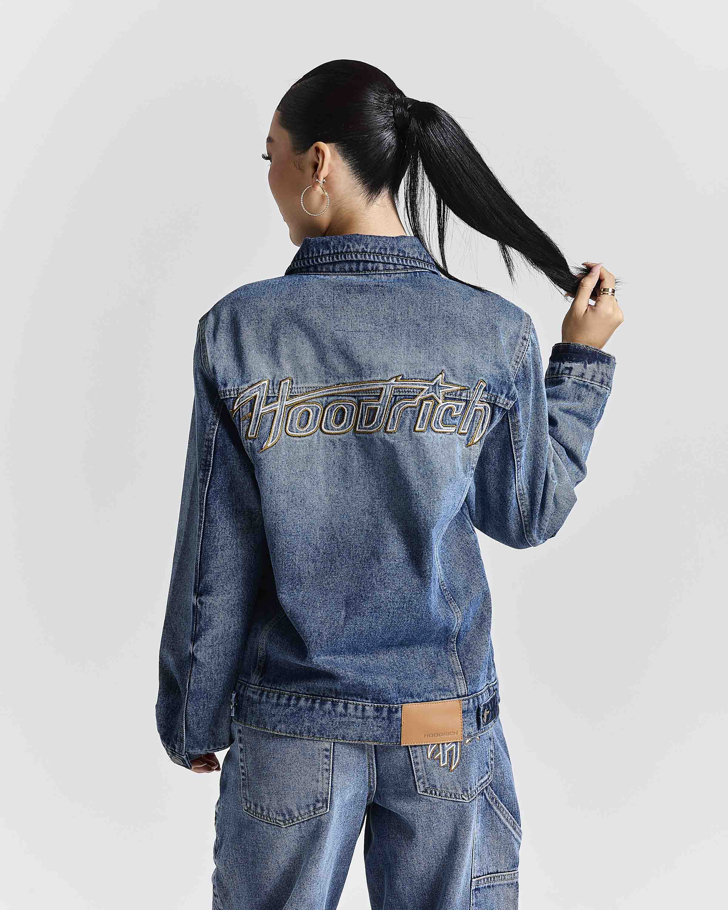 A woman shown from behind wearing a blue denim jacket with 'Hoodrich' embroidered across the back, paired with matching blue jeans, against a light gray studio background.
