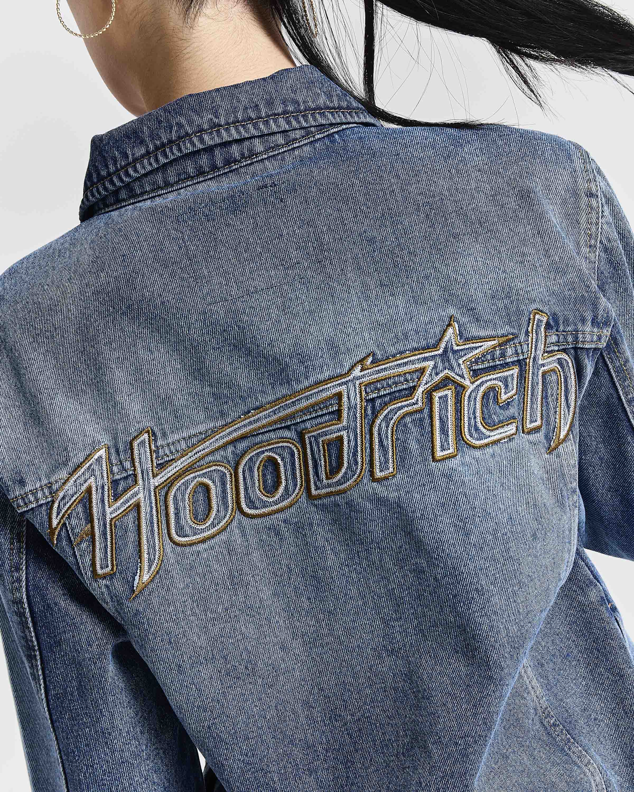 Close-up back view of a person wearing a blue denim jacket with large metallic embroidered 'Hoodrich' lettering across the upper back.