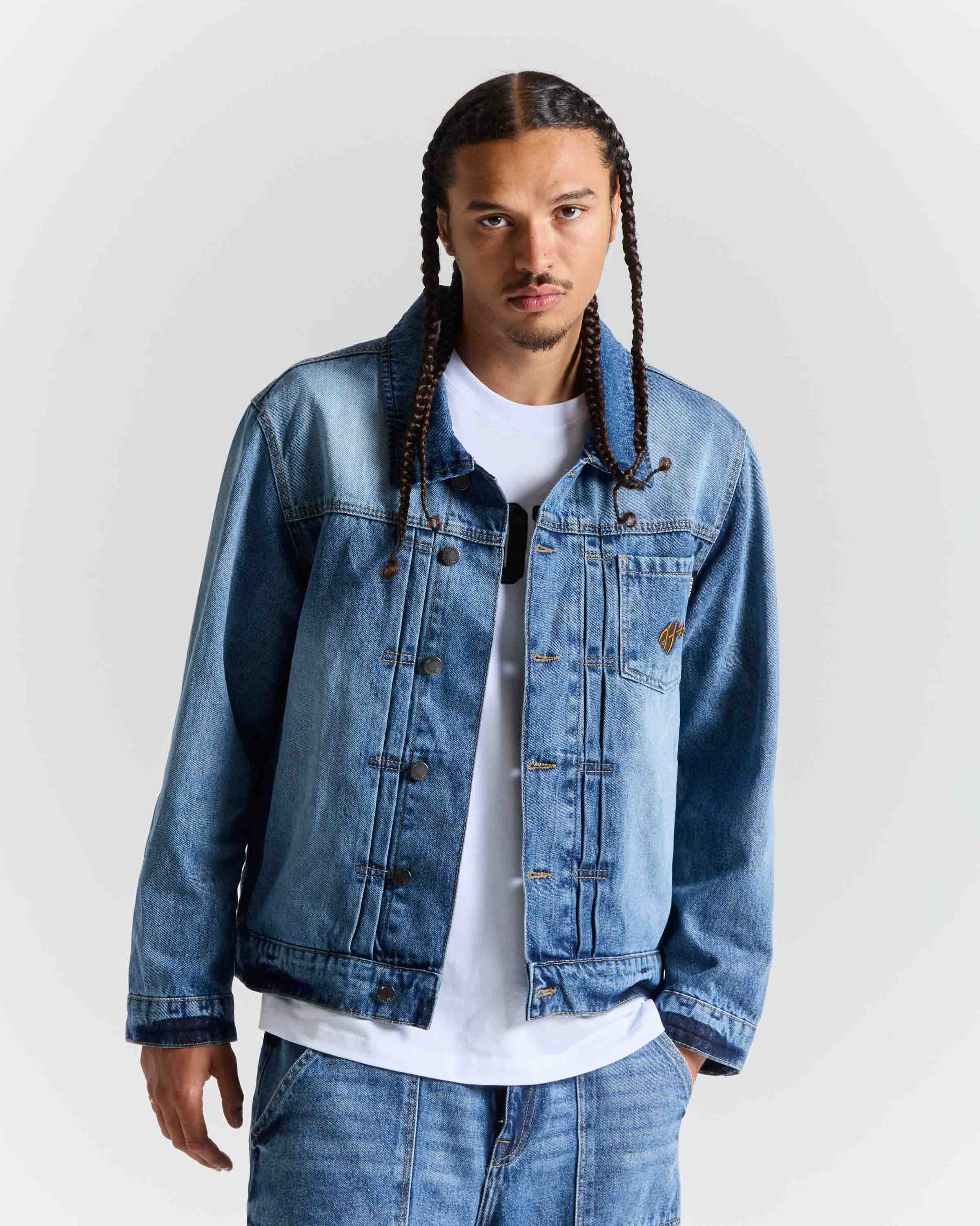 A man with long braids wearing a light blue denim trucker jacket over a white t-shirt and matching blue jeans, photographed against a light gray studio background.