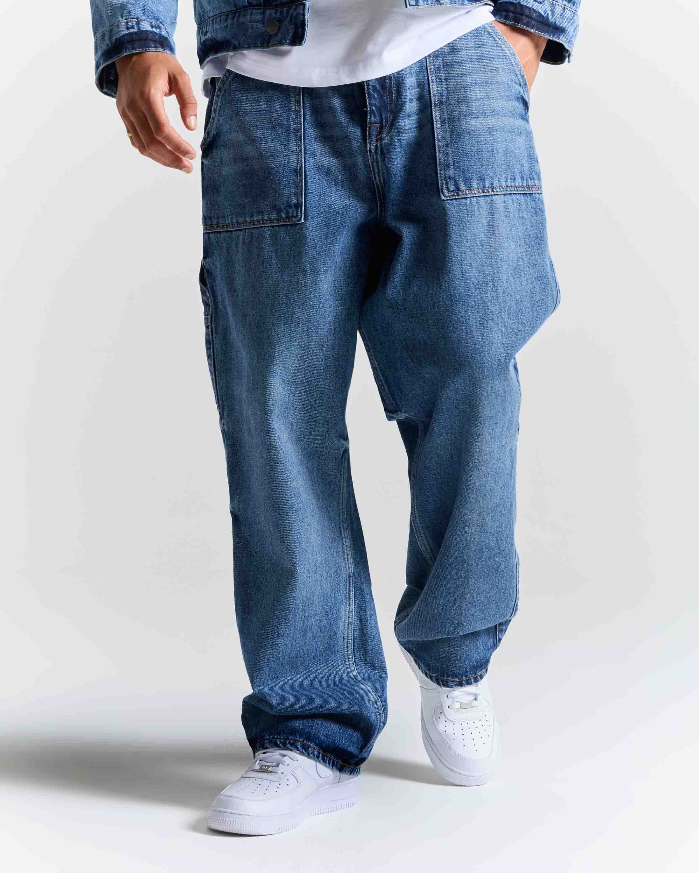 A person wearing loose-fit medium-wash blue denim jeans with a white t-shirt and white sneakers, shown from the waist down against a light gray studio background.