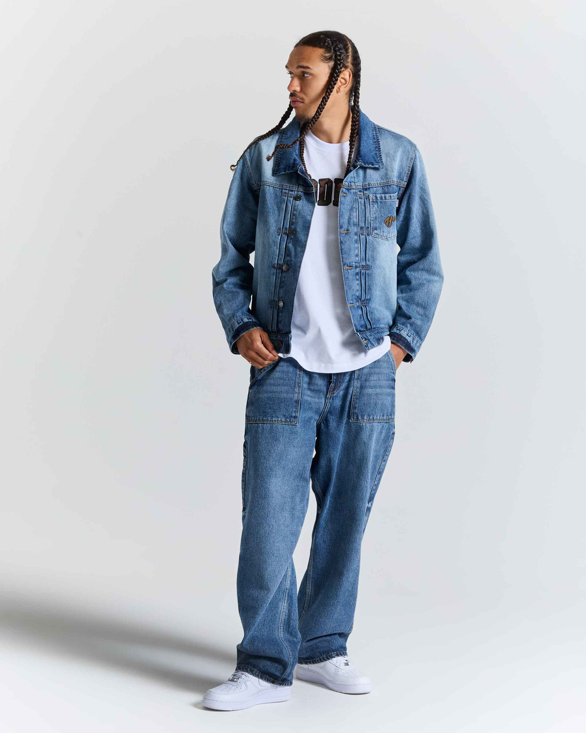 A man modeling a light blue denim jacket and matching wide-leg jeans over a white t-shirt and white sneakers, standing against a light gray studio background.