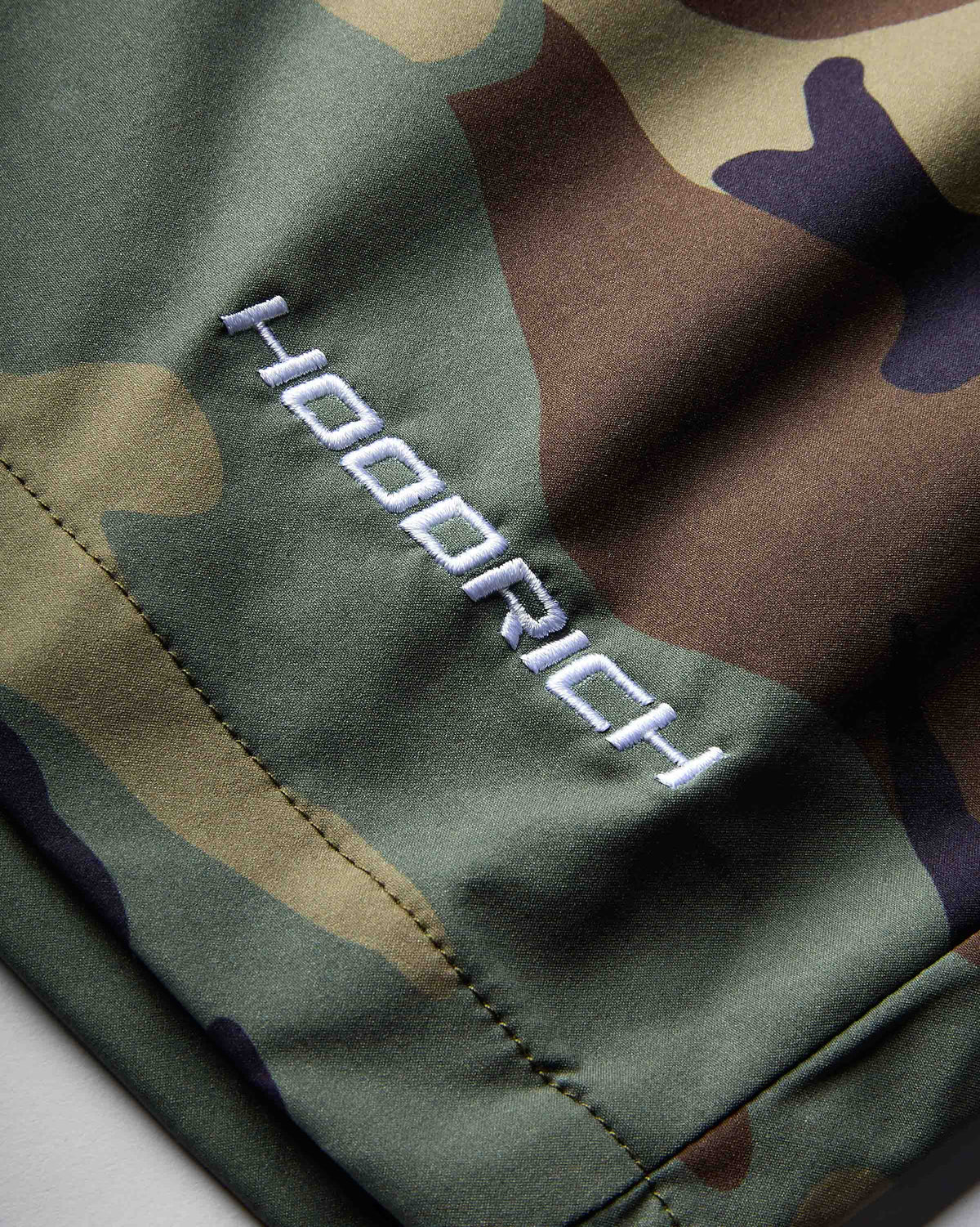 Close-up of a camouflage-patterned garment with a white embroidered 'HOODRICH' logo on the side, showing olive green, brown, tan, and dark purple camo colors.