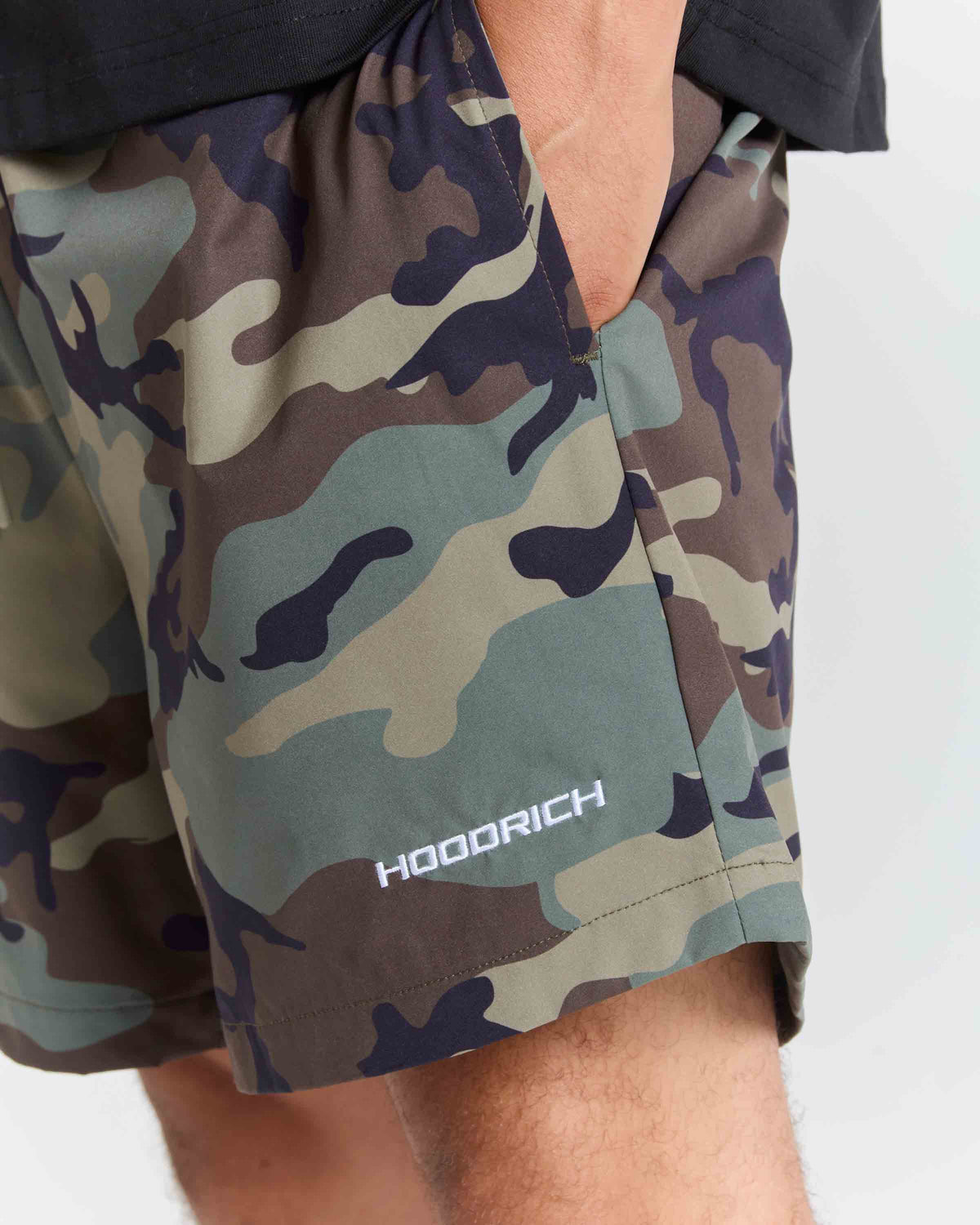 Close-up of a person wearing camouflage shorts with a visible HOODRICH logo near the hem, against a light gray background.