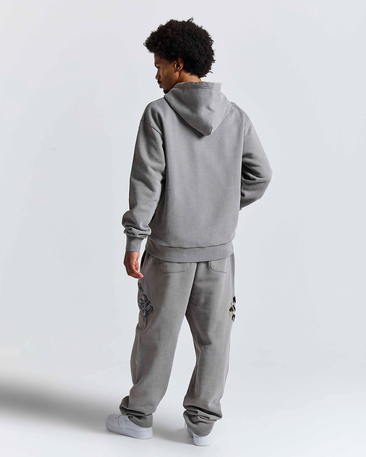 A man wearing a matching gray hoodie and sweatpants set, shown from a rear three-quarter view against a plain light gray studio background.