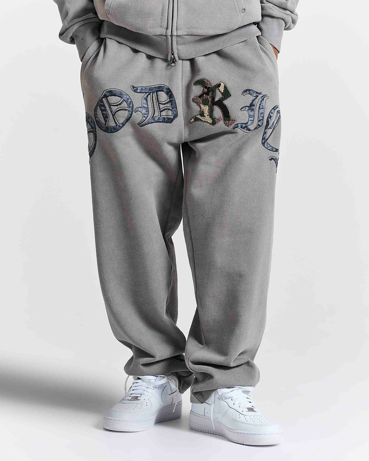 A person wearing gray sweatpants with large decorative embroidered lettering on the legs, paired with white sneakers, shown from the waist down against a light gray studio background.