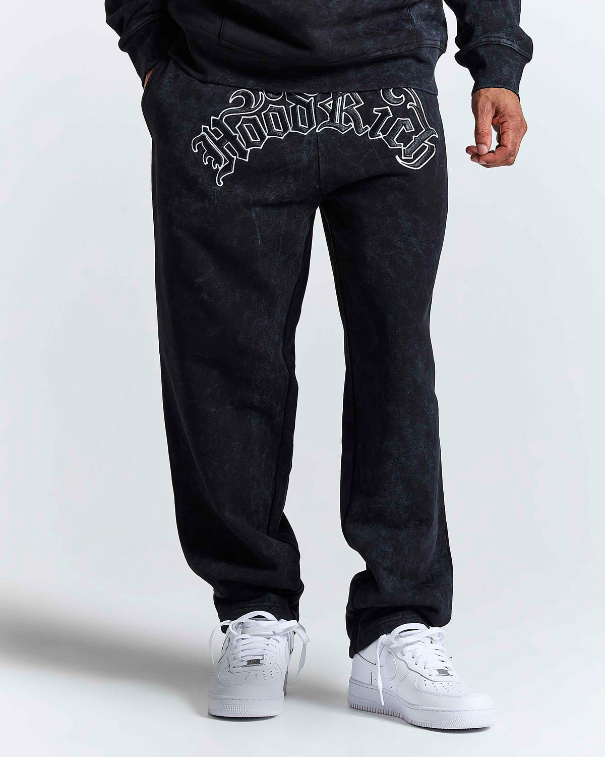 A person wearing black acid-wash sweatpants with a white gothic-style 'Hoodrich' graphic on the back pocket, paired with white sneakers, shown from the waist down against a light gray studio background.