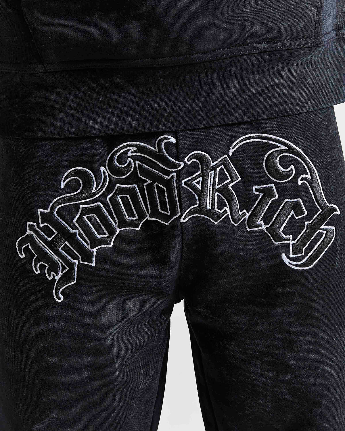 Close-up of black velour sweatpants featuring a large white embroidered gothic-style 'Hoodrich' graphic across the back pocket.