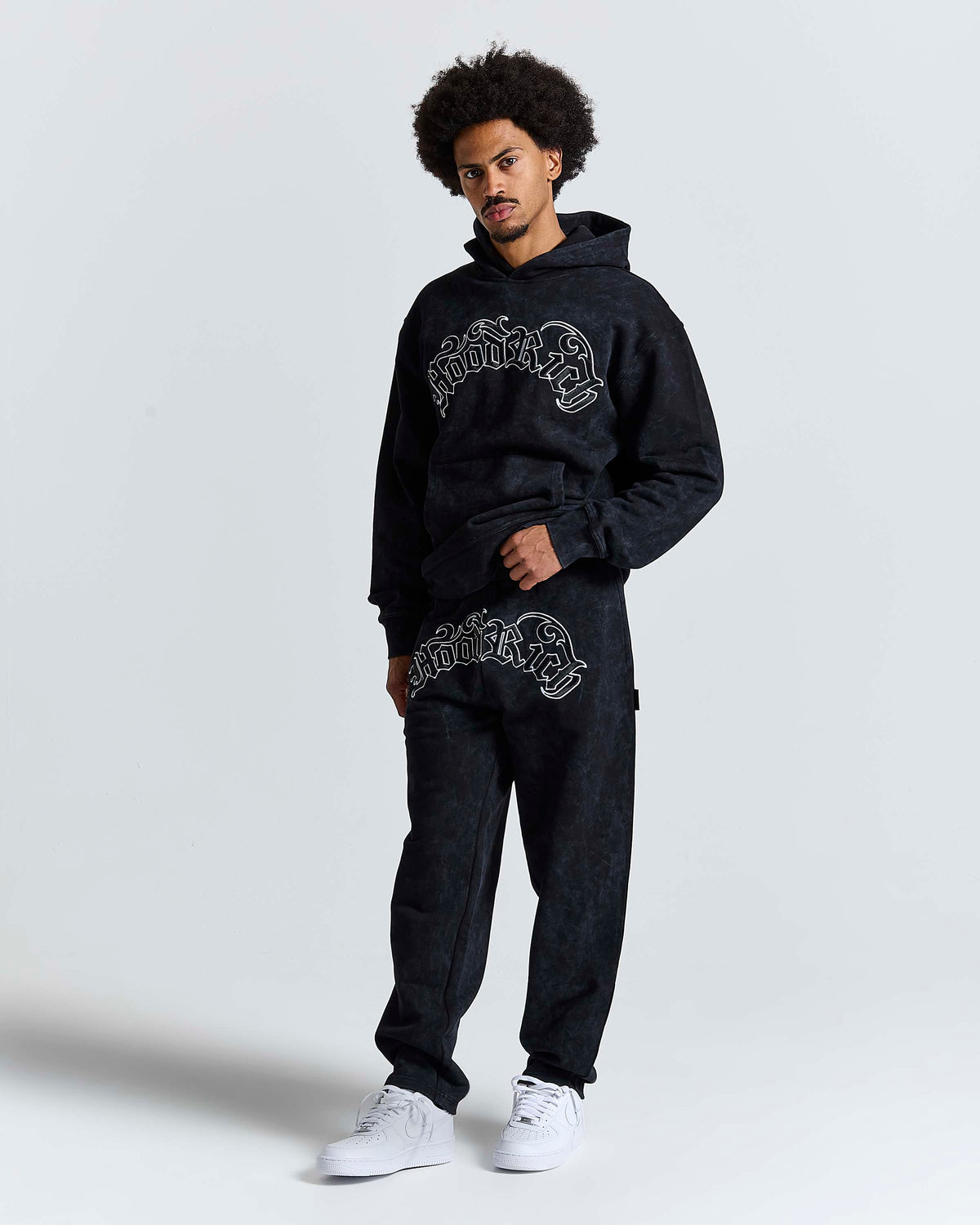 A man modeling a black hooded sweatshirt and matching jogger pants set with white graphic text across the chest and upper thigh, paired with white sneakers, against a light gray studio background.
