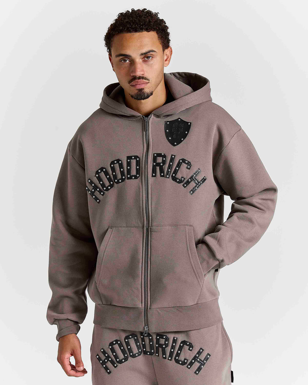 Displays a model wearing the Falcon Black 'hoodrich' branded Varso Studs Oversized Zip Through Hoodie
