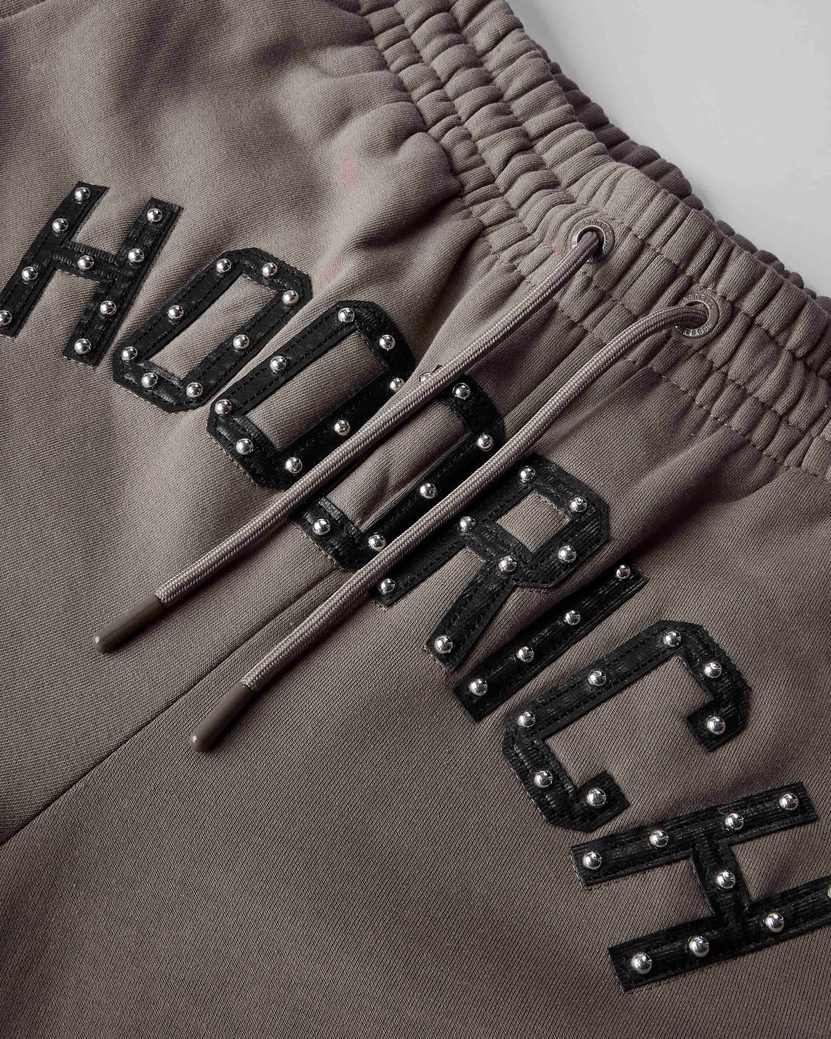 Close-up of taupe-brown sweatpants featuring a large black embroidered 'HOODRICH' text across the leg, embellished with silver studs, and a matching brown drawstring at the elastic waistband.