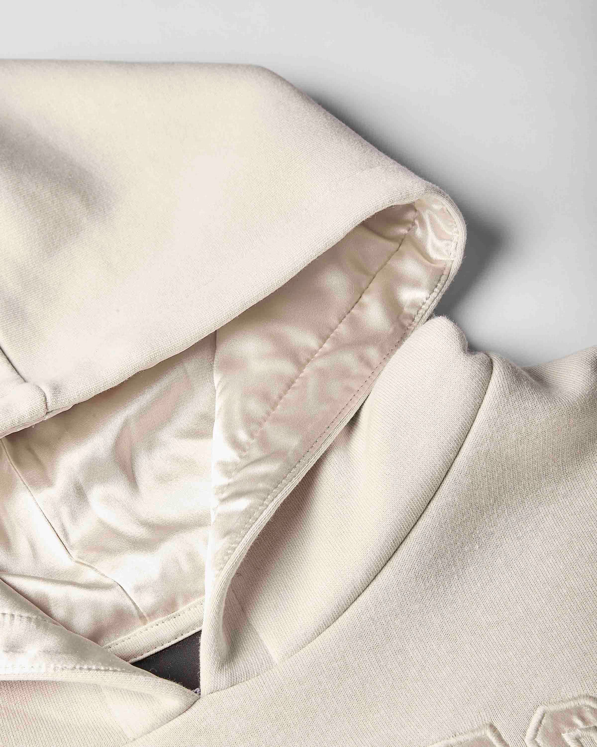 Close-up of a cream-colored hoodie showing the interior satin lining of the hood and collar, featuring a smooth satin-like inner fabric and a textured knit outer fabric.