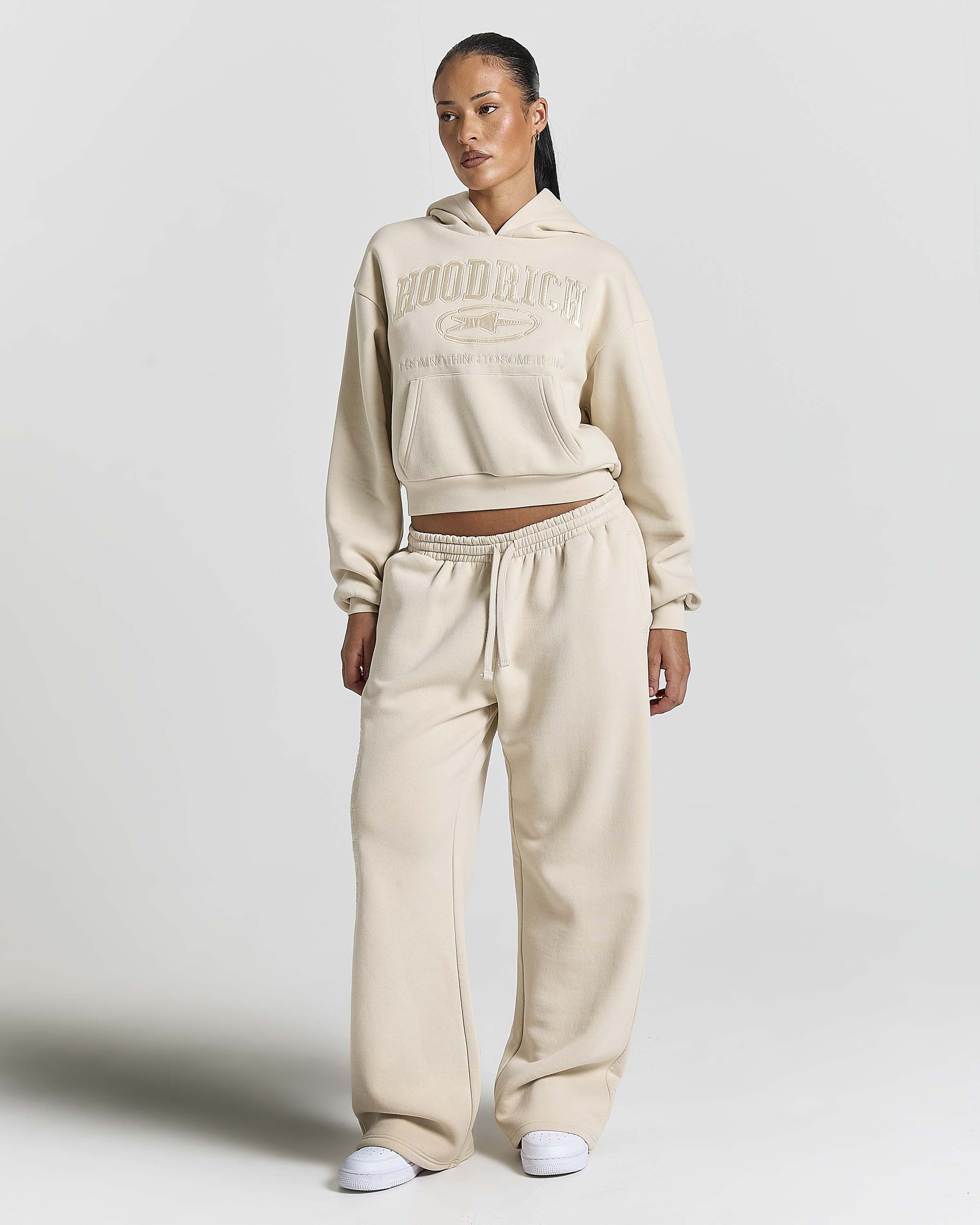 A model wearing a matching beige cropped hoodie and wide-leg sweatpants set with white sneakers, standing against a light gray studio background.