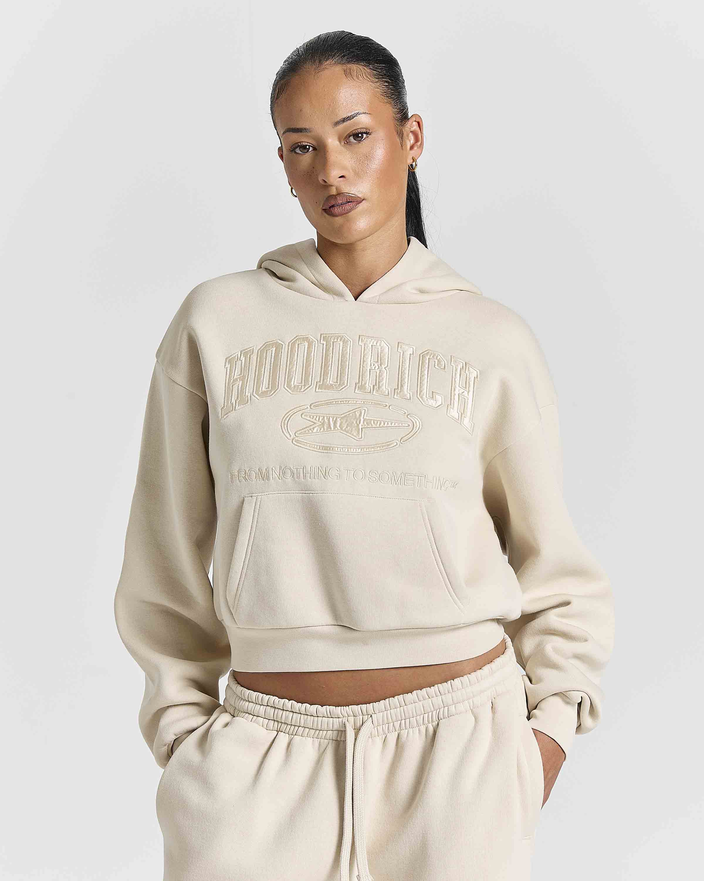A model wearing a cream-colored cropped hoodie with 'HOODRICH' embossed on the chest, paired with matching jogger pants, against a light gray studio background.