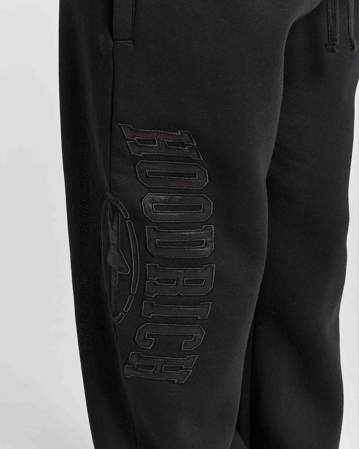 Close-up of black sweatpants featuring a large tonal black-on-black 'HOODRICH' logo and circular emblem on the left leg, shown against a light gray background.