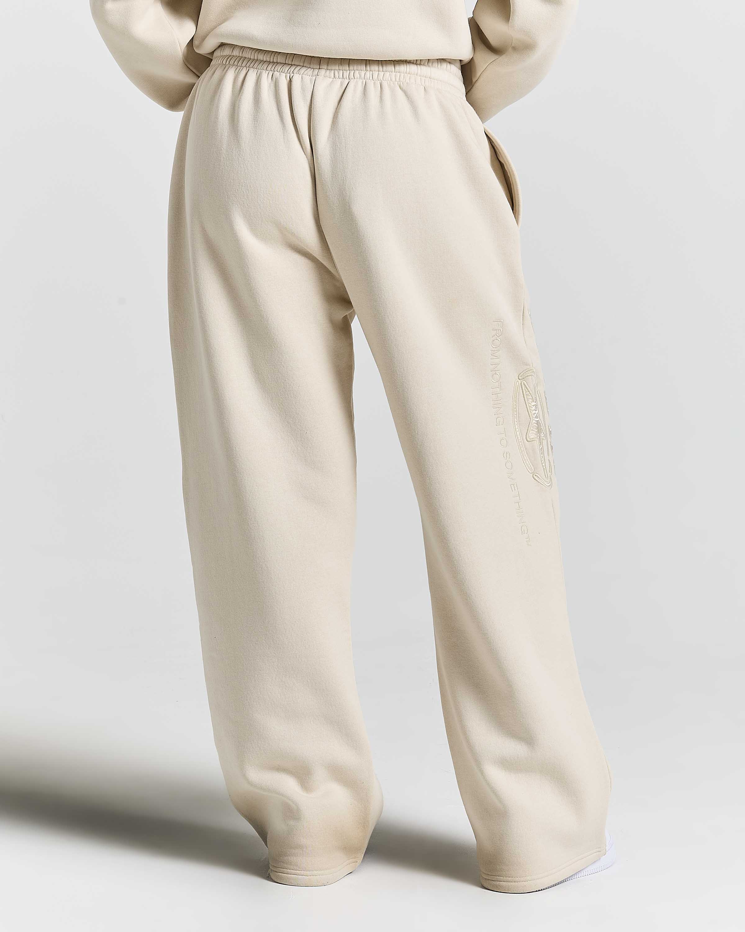 A person shown from the back wearing loose, wide-leg beige sweatpants with an elastic waistband and a tonal embroidered graphic on the lower right leg, against a light gray studio background.